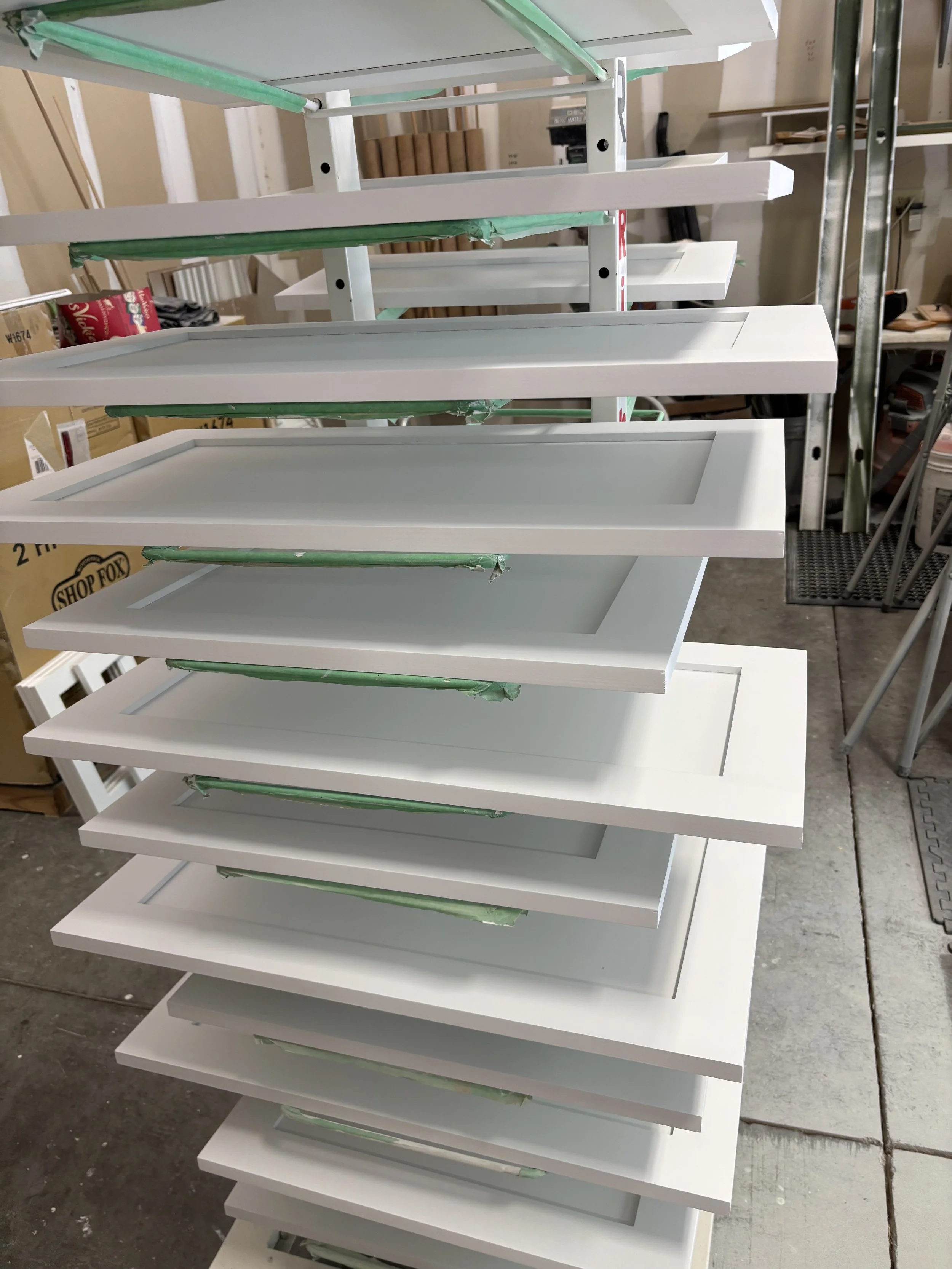 Stacked white shelving units in a workshop or storage area, with green protective padding on some shelves, and various boxes and tools visible in the background.