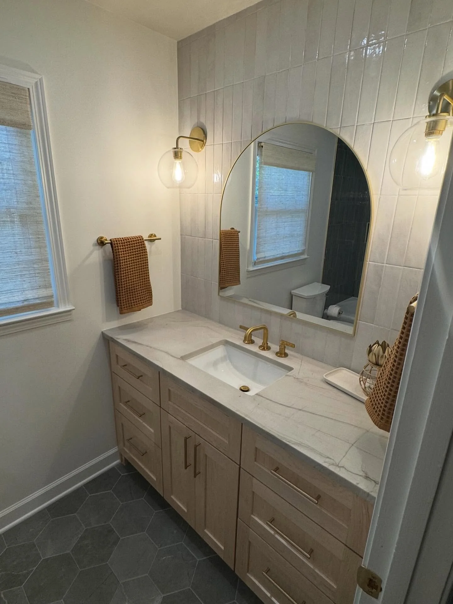 A bathroom vanity with a white marble countertop, a built-in sink, brass faucet, wooden drawer cabinets, a large mirror, wall-mounted brass and glass light fixtures, a window with a beige shade, and dark hexagonal tile flooring.