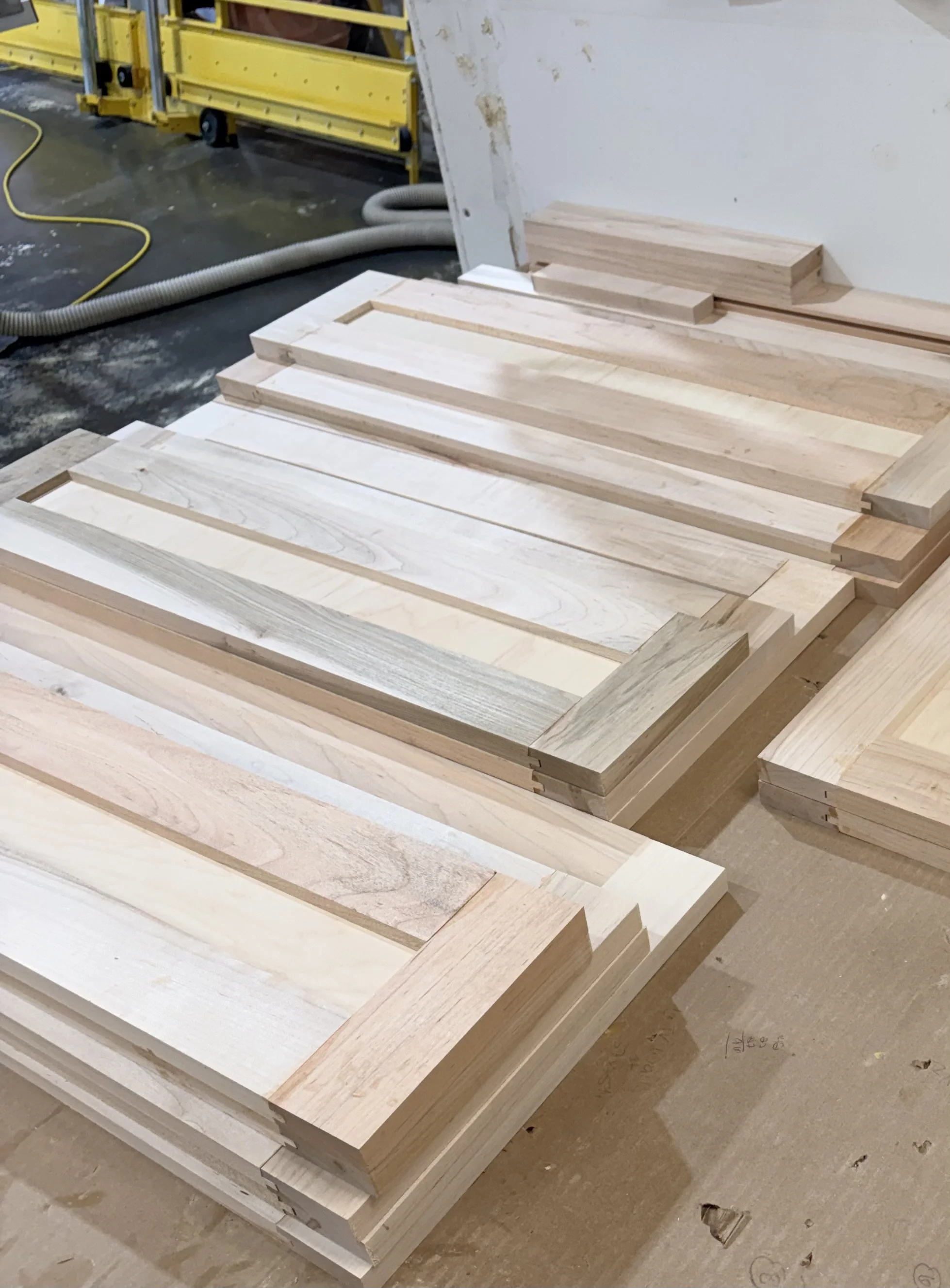 Stacked wooden boards in a workshop with a yellow tool cart and a flexible hose in the background.