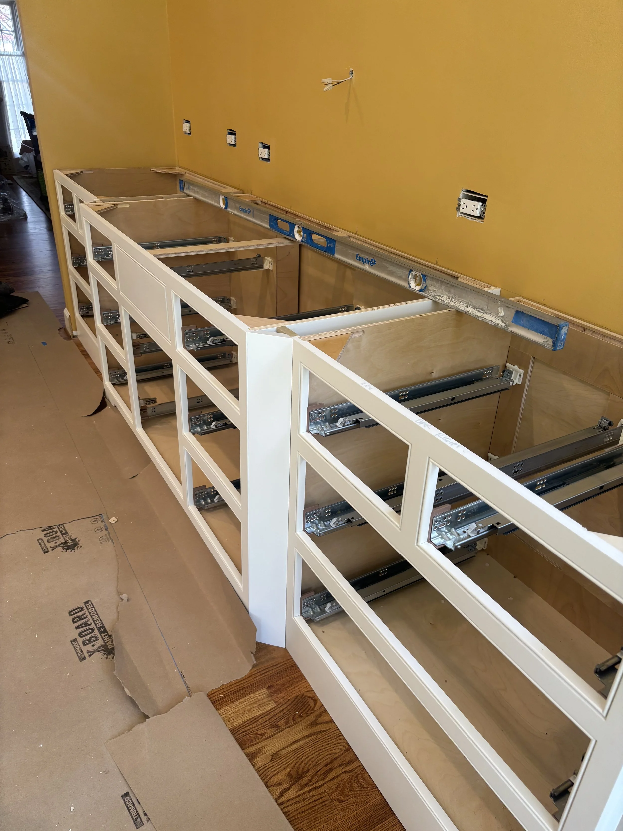Kitchen cabinet frame under construction with white trim, mounting hardware, and wall electrical outlets visible. Yellow wall in background, hardwood floor to the right, work supplies nearby.