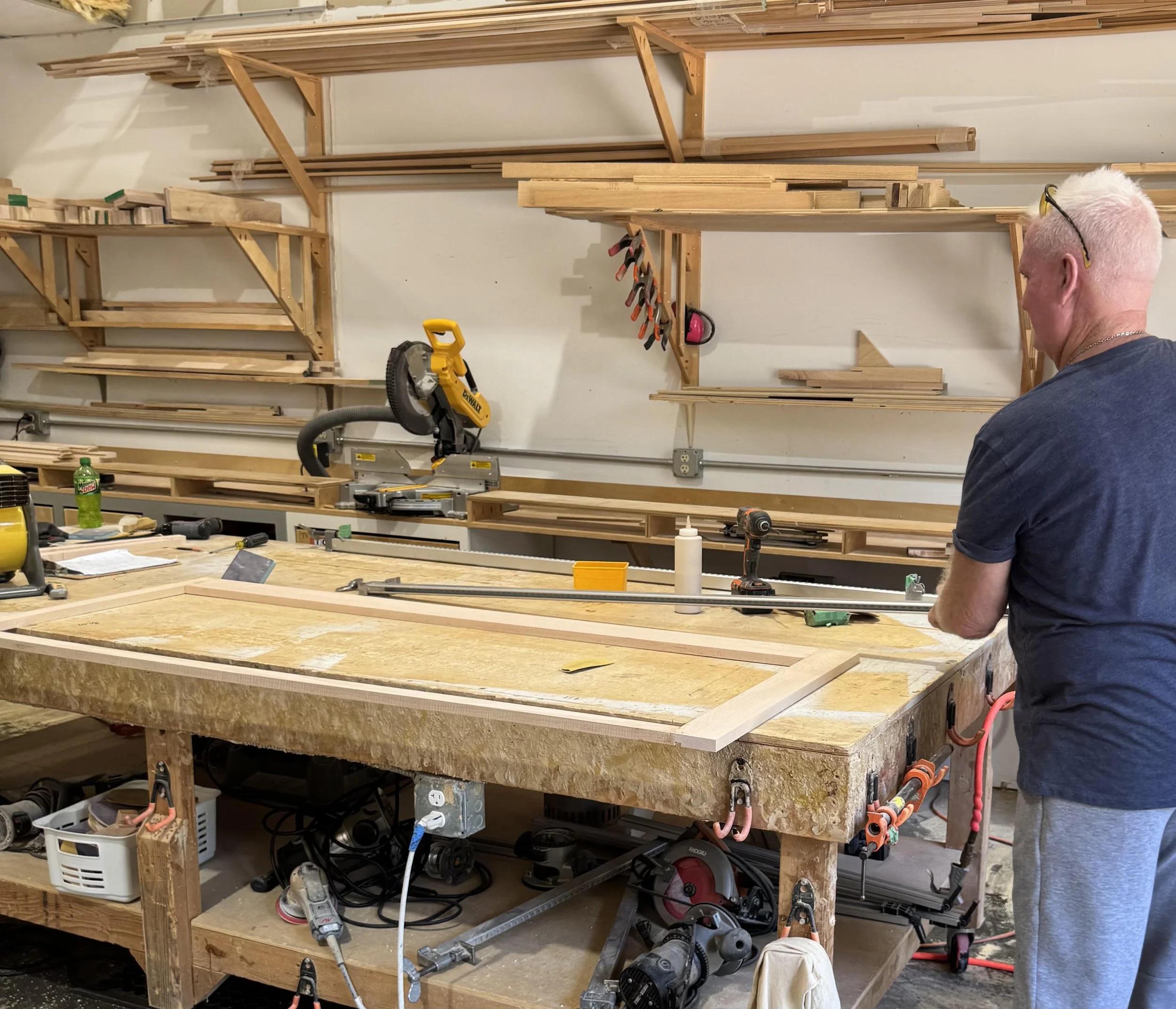 Workshop with wooden shelves and workbench filled with tools and woodworking supplies, a person wearing glasses and a dark shirt facing away.