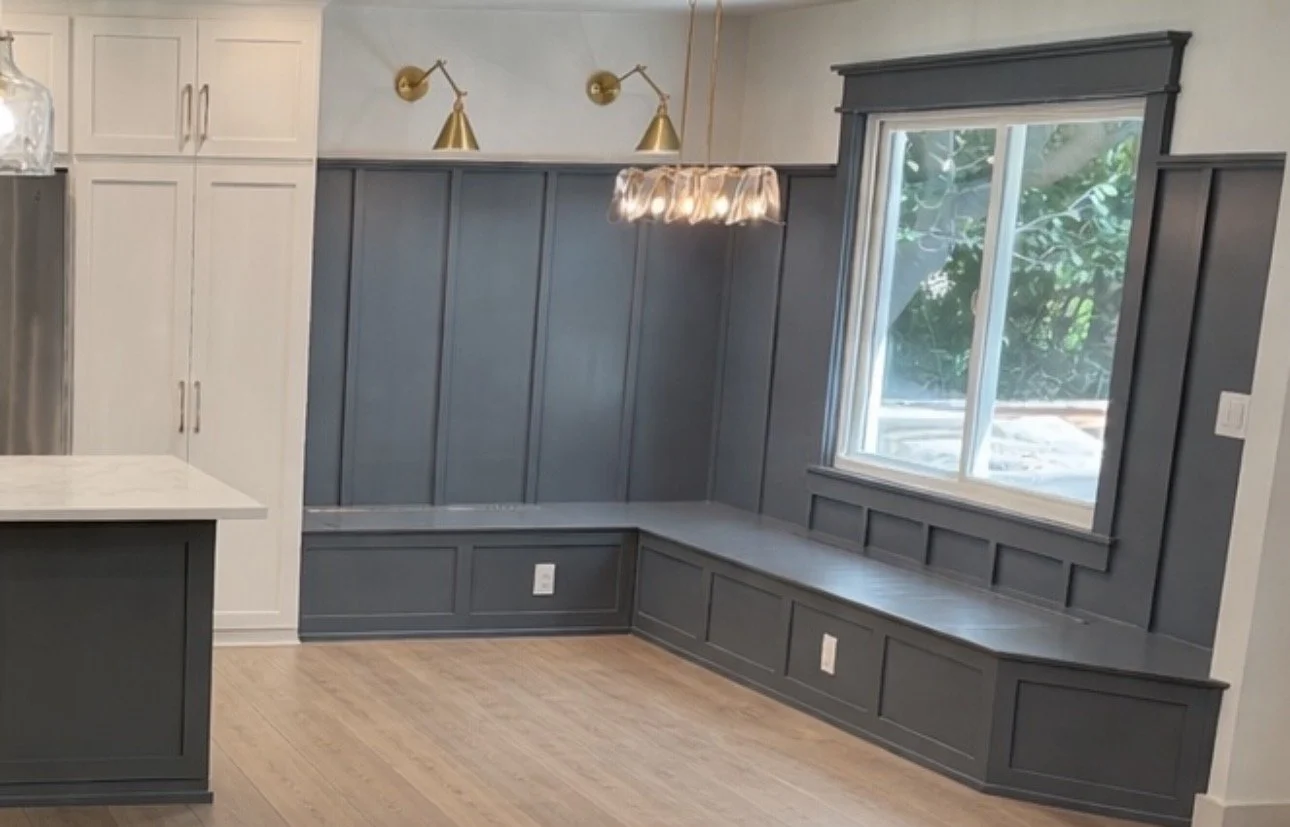 Modern kitchen corner with gray wainscoting, a large window with black trim, a pendant light, two gold wall sconces, white upper cabinets, and light hardwood flooring.