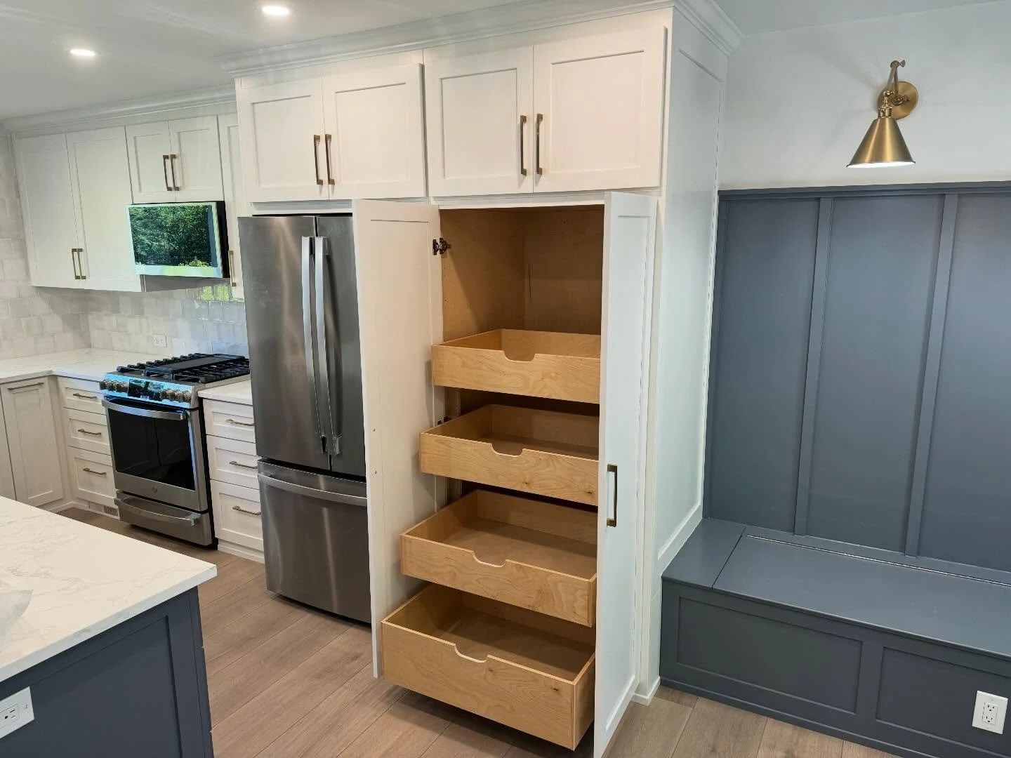 Kitchen with white cabinets, stainless steel refrigerator, oven, and microwave, wood flooring, and a dark gray built-in bench with a wall sconce.