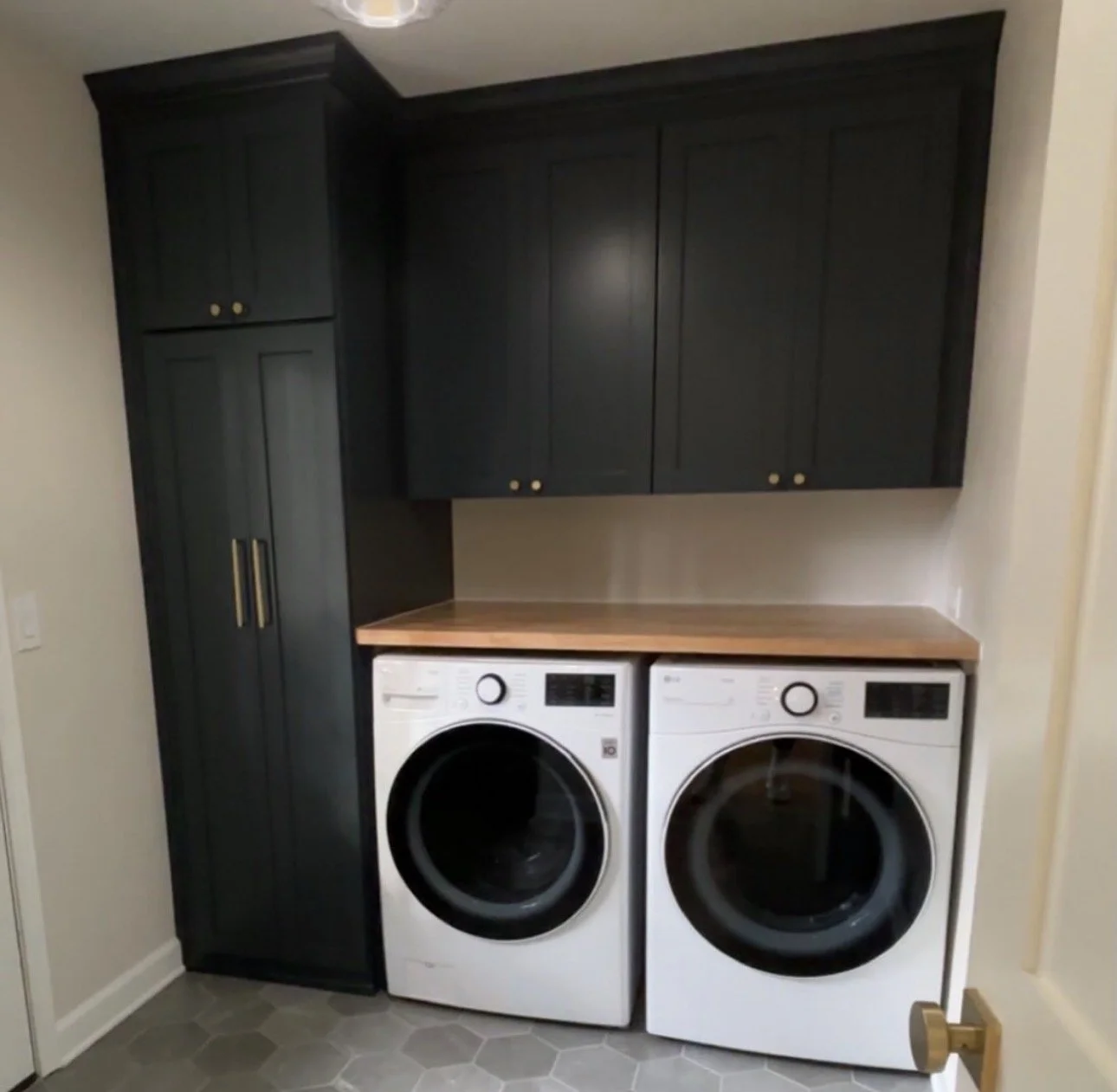 A laundry room with black upper cabinets, a wooden countertop, and a beige wall. There are two front-loading washing machines underneath the countertop, with hexagonal gray floor tiles.
