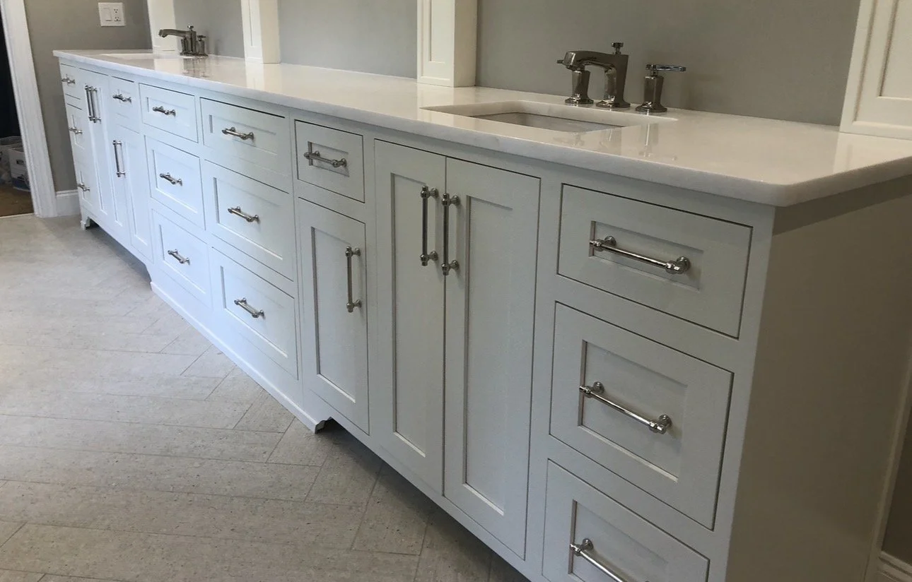 White bathroom vanity with multiple drawers and cabinets, silver handles, and a marble countertop with two built-in sinks and silver faucets.