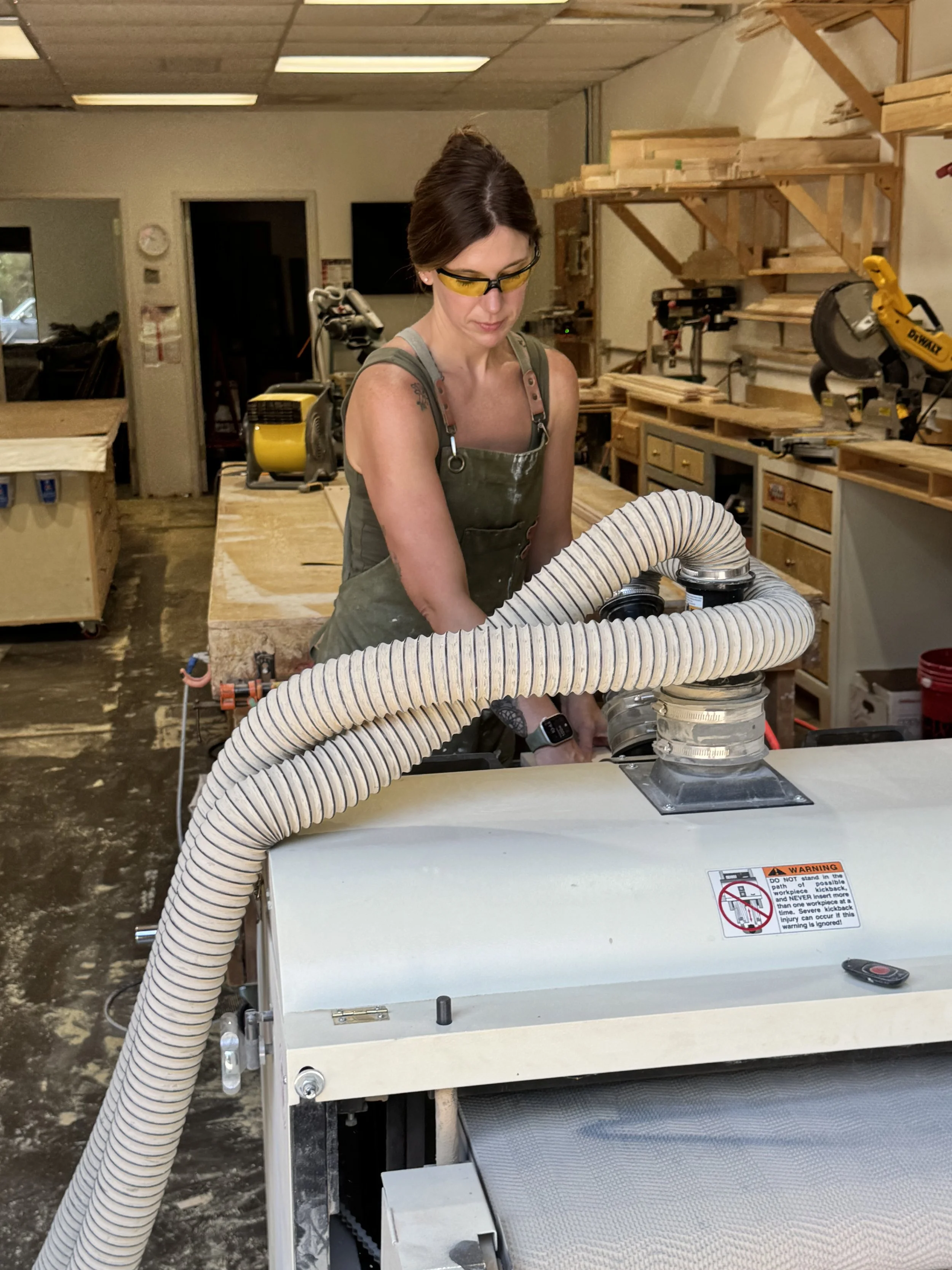 A woman working in a woodshop wearing safety glasses and an apron, with woodworking tools and shelves in the background, and connected to a machine with a large flexible hose.