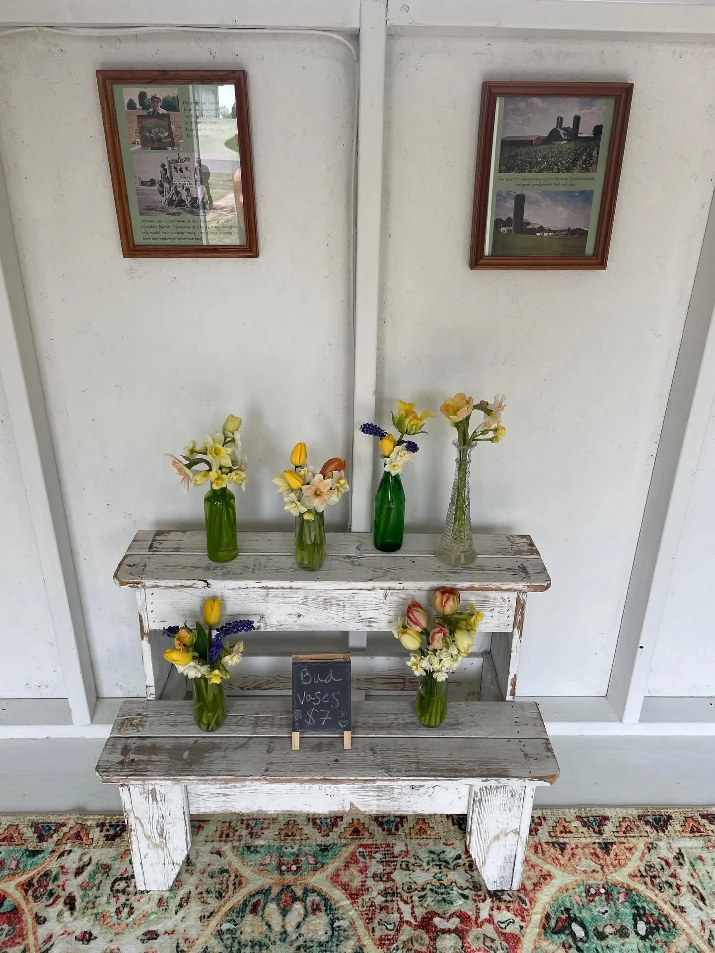 White wooden stand with three shelves displaying various colorful bud vases with flowers, a small chalkboard sign, and framed wall art with text and images of farm scenes, on a patterned rug.