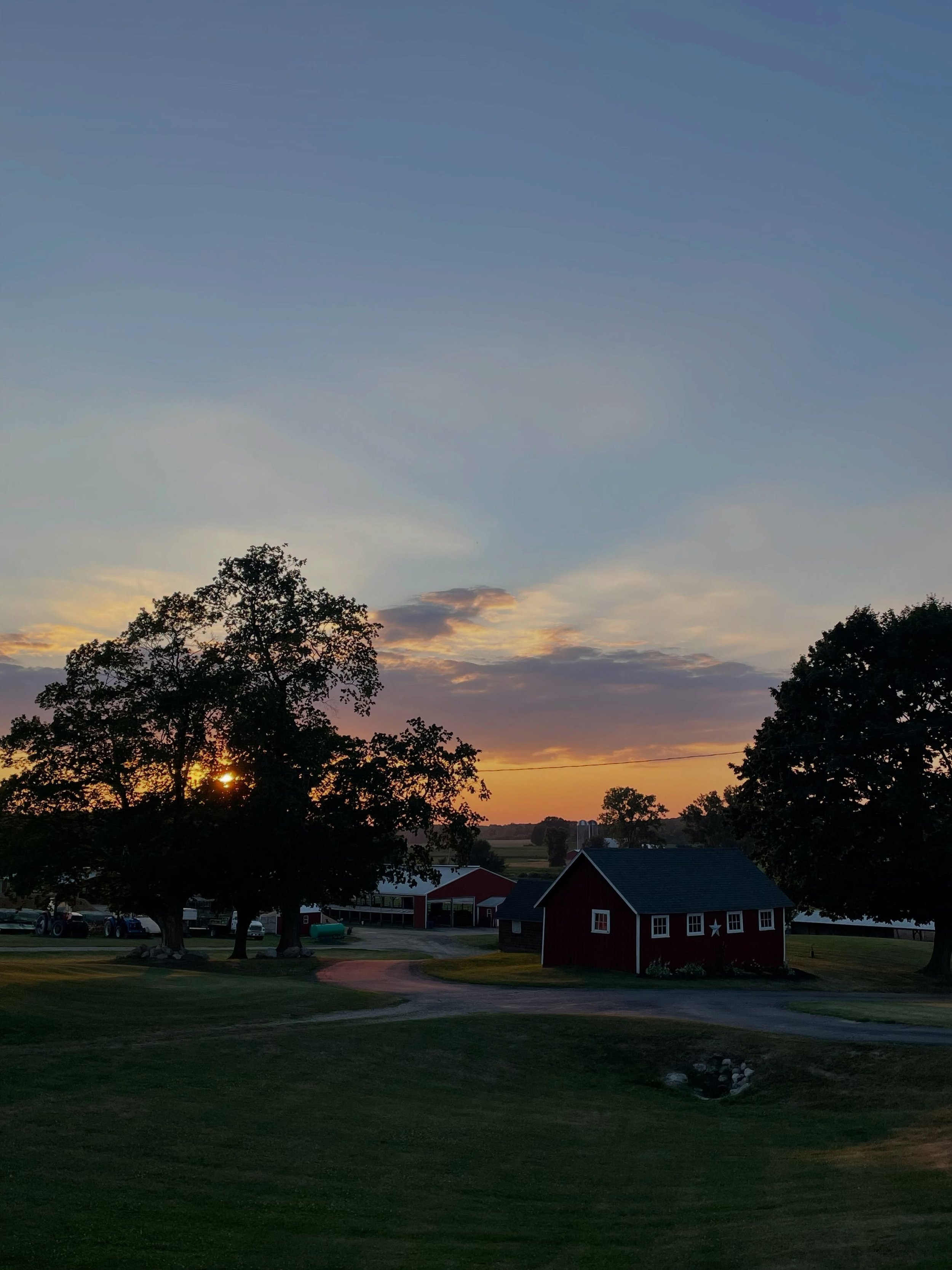 A rural landscape at sunset with trees, a red barn, small buildings, a parking lot, and a sky with clouds and the sun near the horizon.