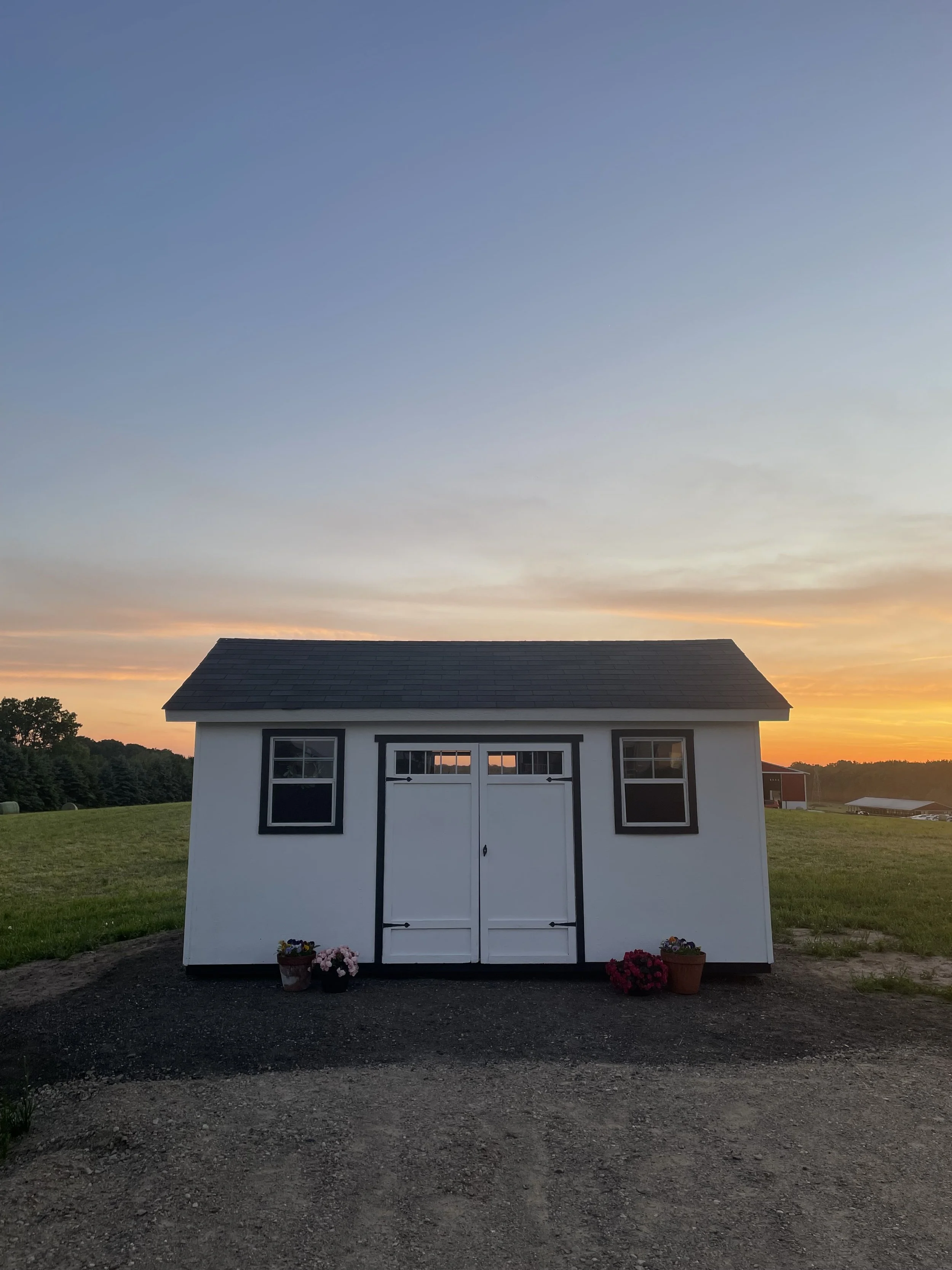 First Bloom Flower Stand during sunset. There are flower pots with flowers in front of the shed, and trees and other buildings are visible in the background under a colorful sky.