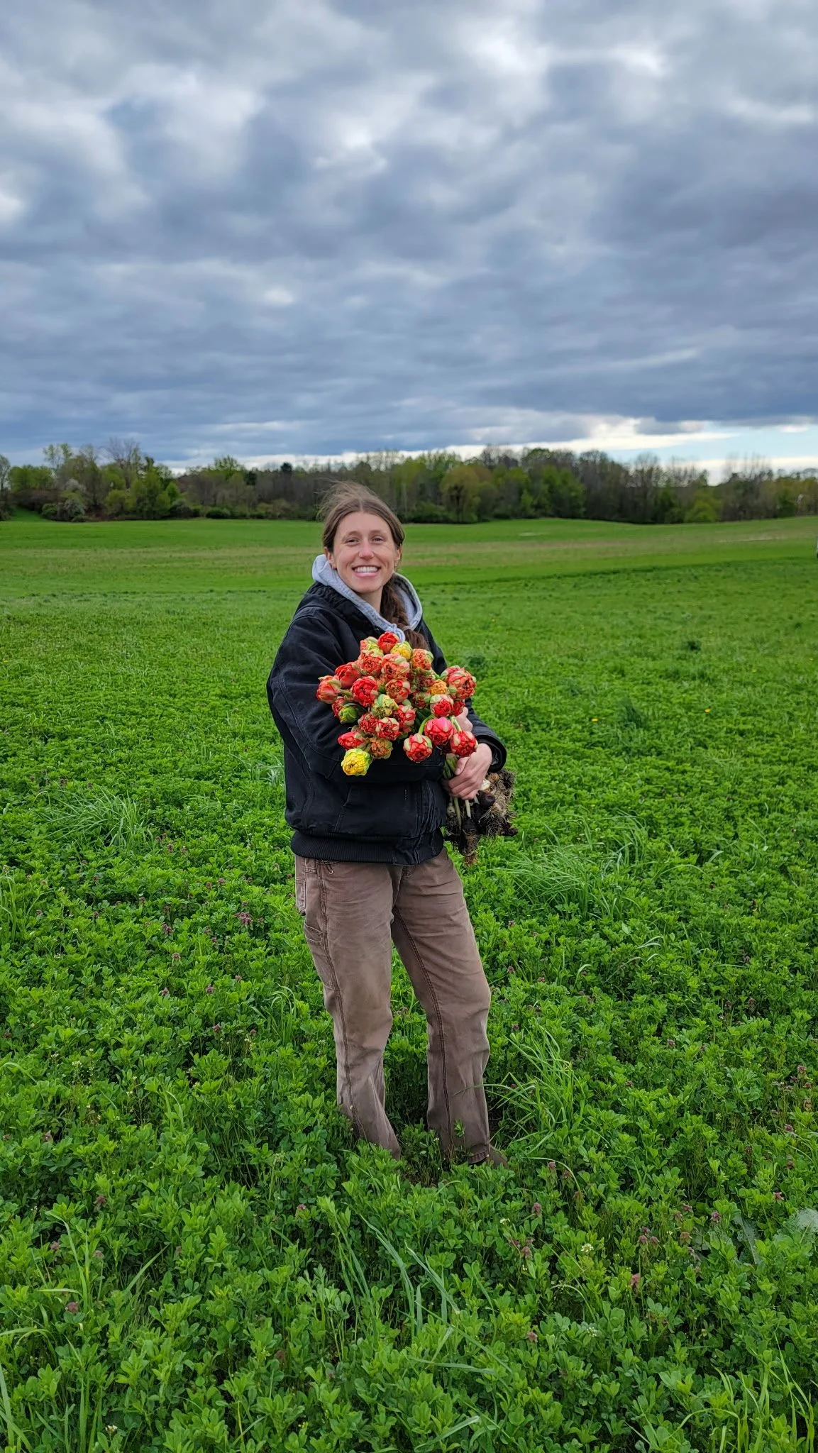 Morgan, farmer at First Bloom Farm, smiling and holding a large bouquet of flowers, standing in a green field under a cloudy sky.