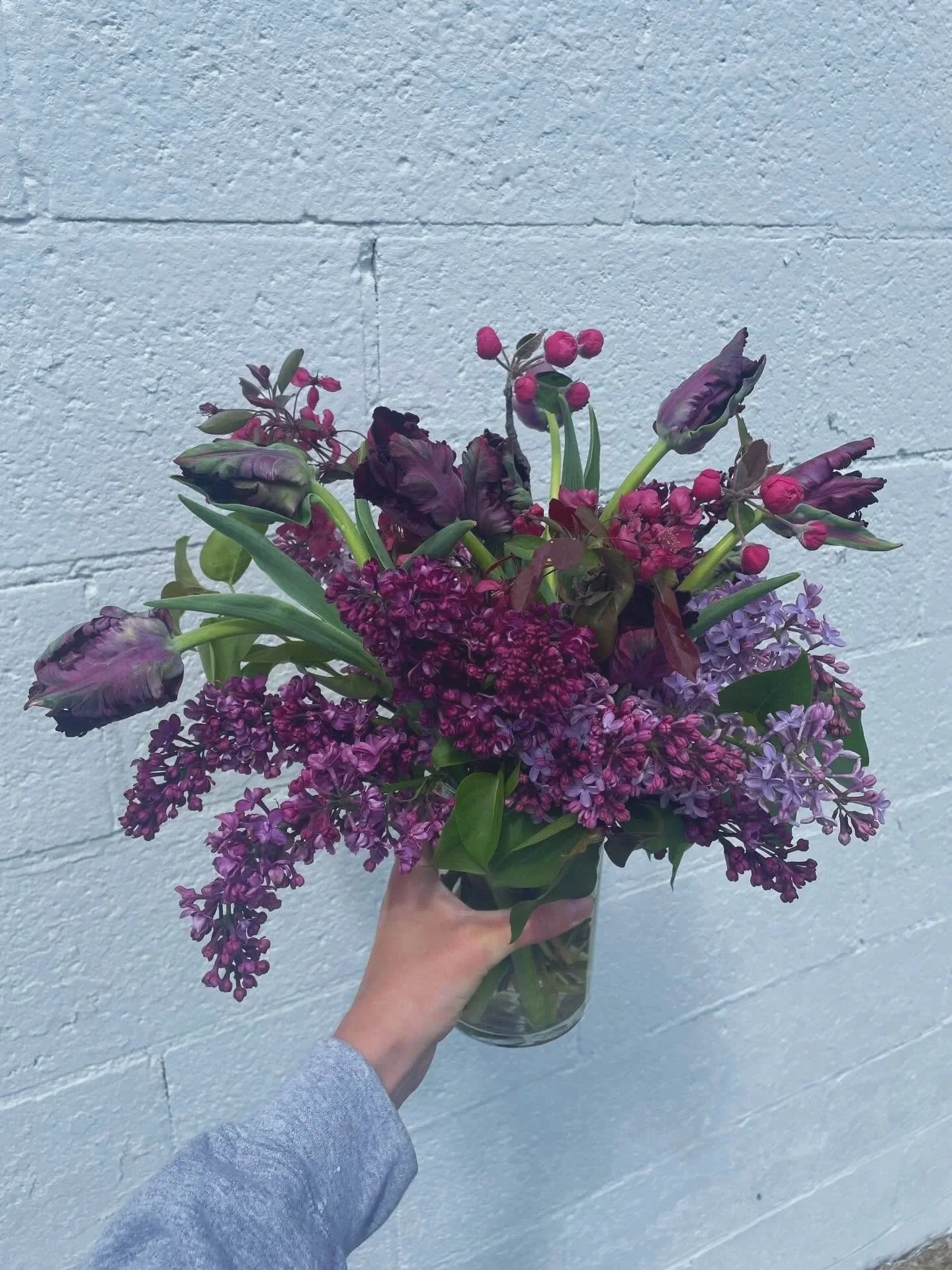 A hand holding a clear glass vase with a bouquet of purple and pink flowers in front of a white brick wall.
