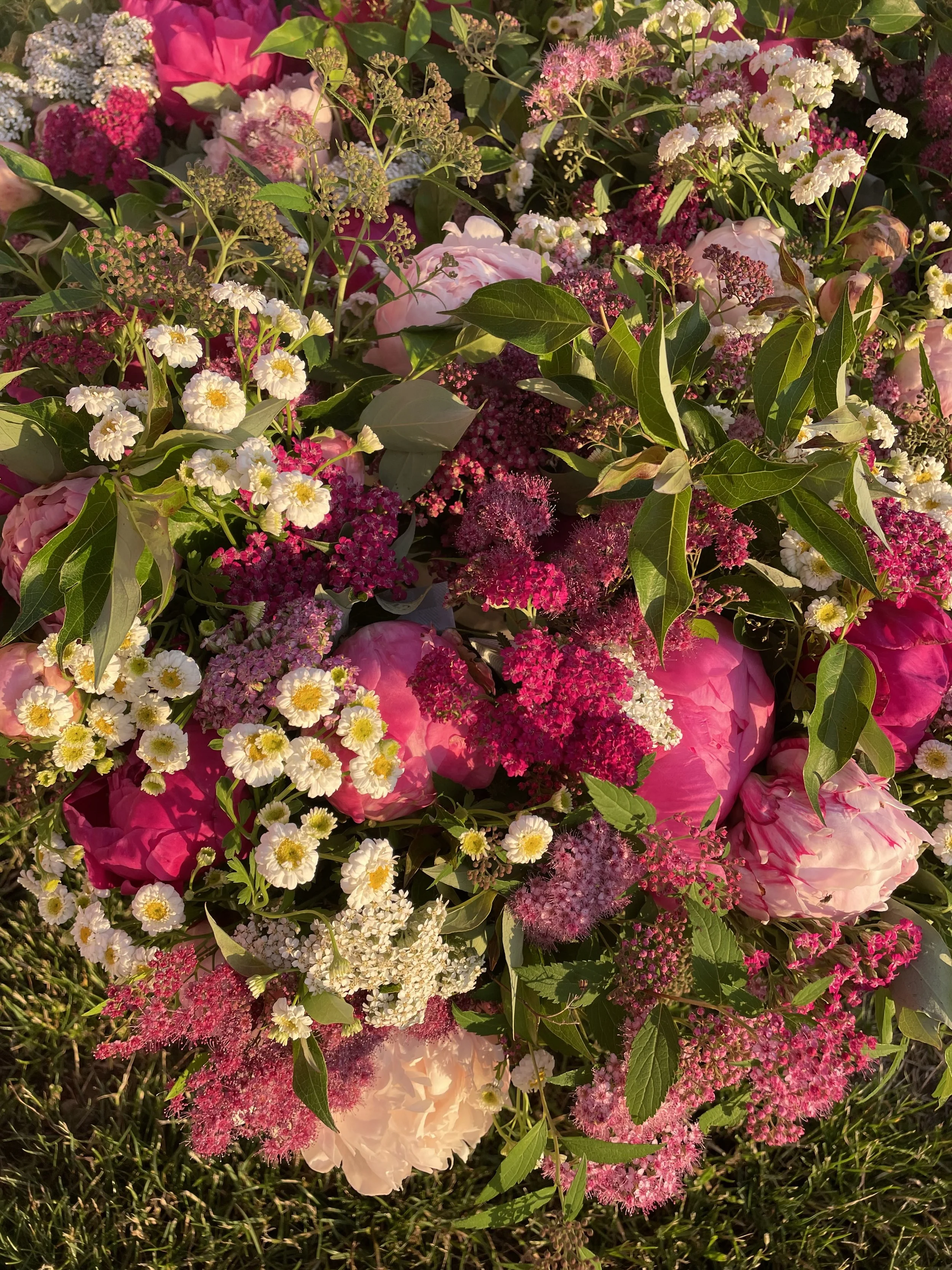 A colorful flower arrangement with pink, white, and purple flowers, including roses, small daisies, and various other blossoms, surrounded by green leaves and grass.