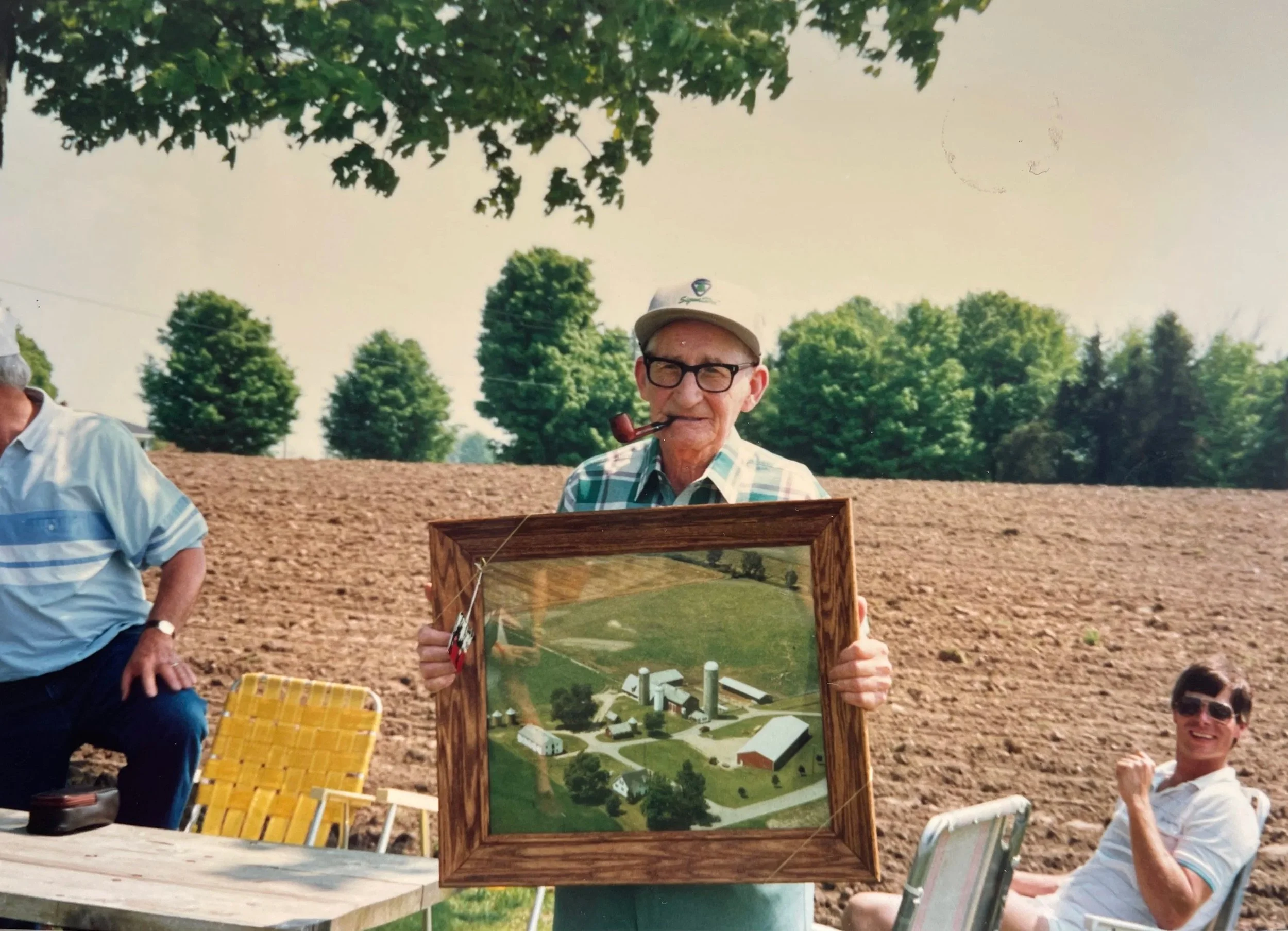 An elderly man with glasses, a cap, and a pipe is holding a framed aerial view picture of a property. There are other people and outdoor furniture in the background, indicating a casual outdoor gathering.