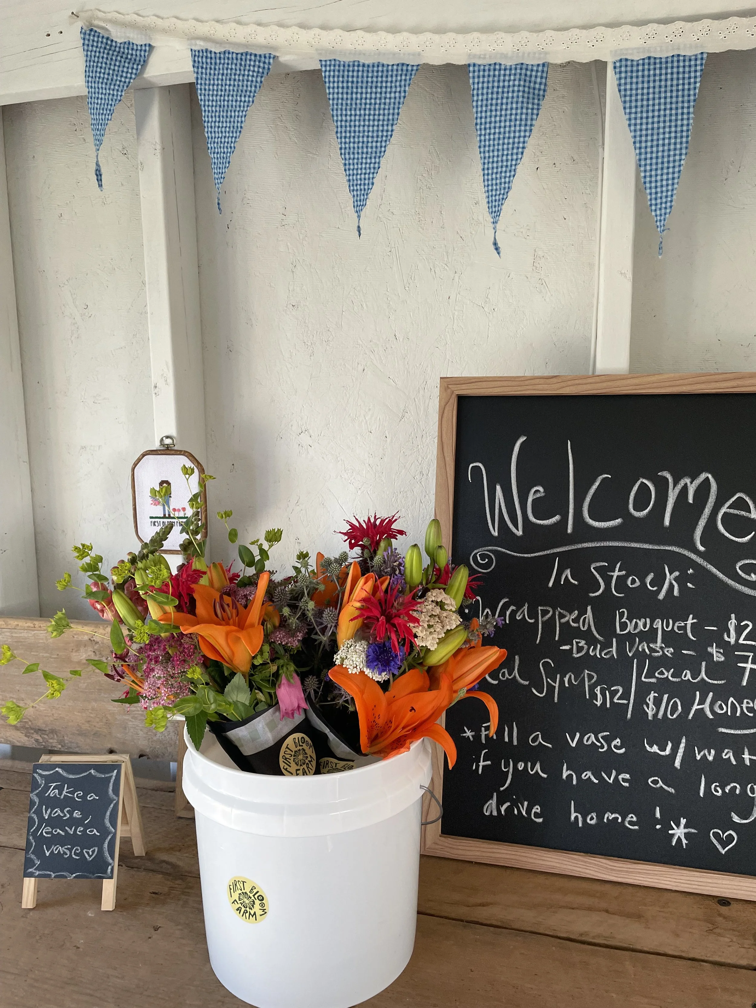 A display of flowers in a white bucket with a logo that reads 'First Bloom Farm', with a chalkboard sign listing flower prices and details, placed next to a smaller chalkboard sign that reads 'Take a Vase, Leave a Vase'.