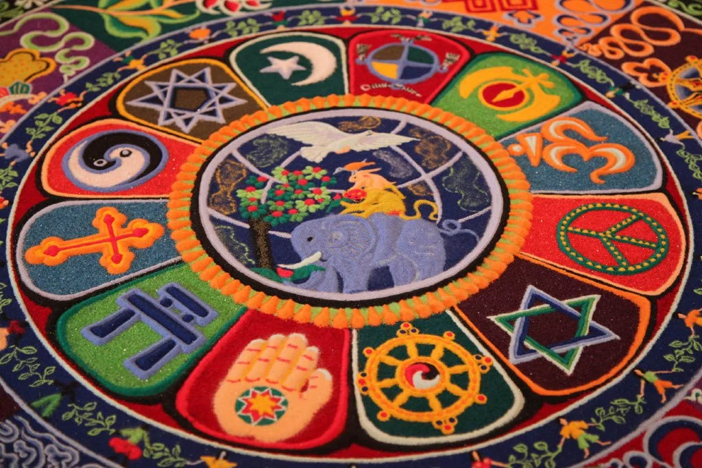 Colorful circular embroidered textile with various symbols and patterns, featuring a central scene with a rabbit, elephant, and bird, surrounded by symbols including a moon, star, peace sign, and others.