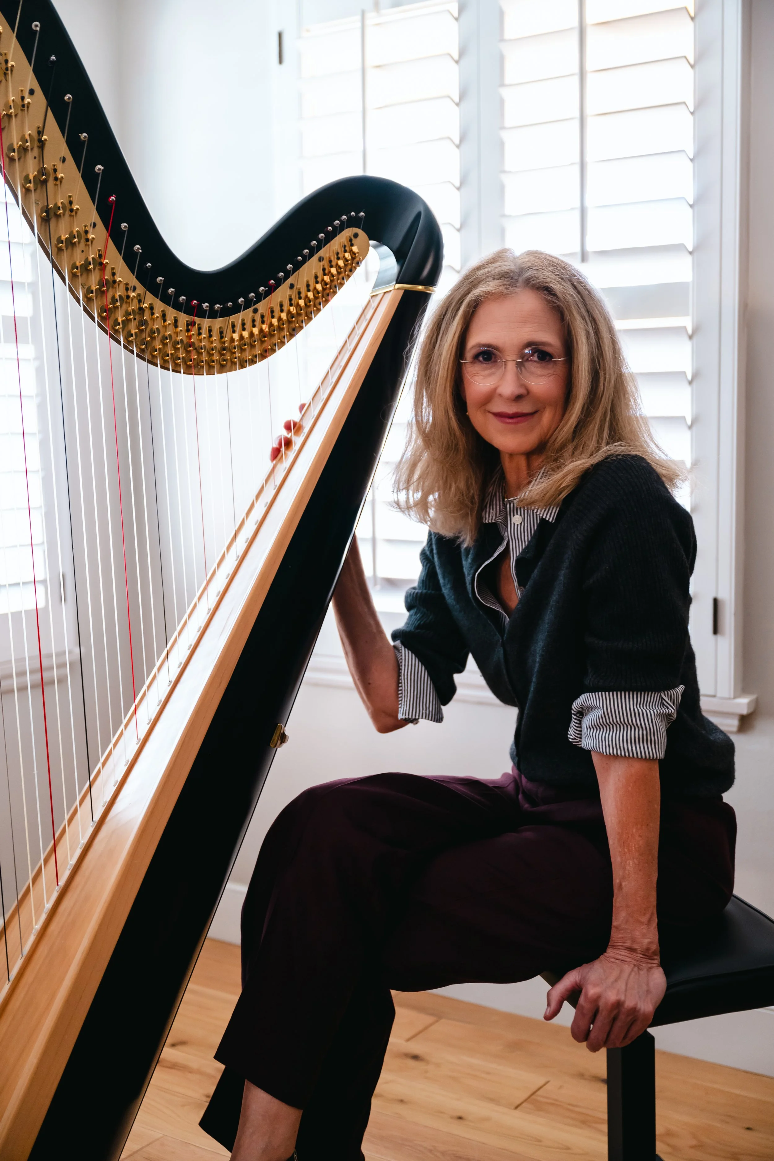 An older woman with glasses and shoulder-length hair sitting next to a harp, smiling at the camera. She is wearing a black cardigan over a striped shirt, and has her arm resting on her knee. The room has white plantation shutters on the window and a wooden floor.