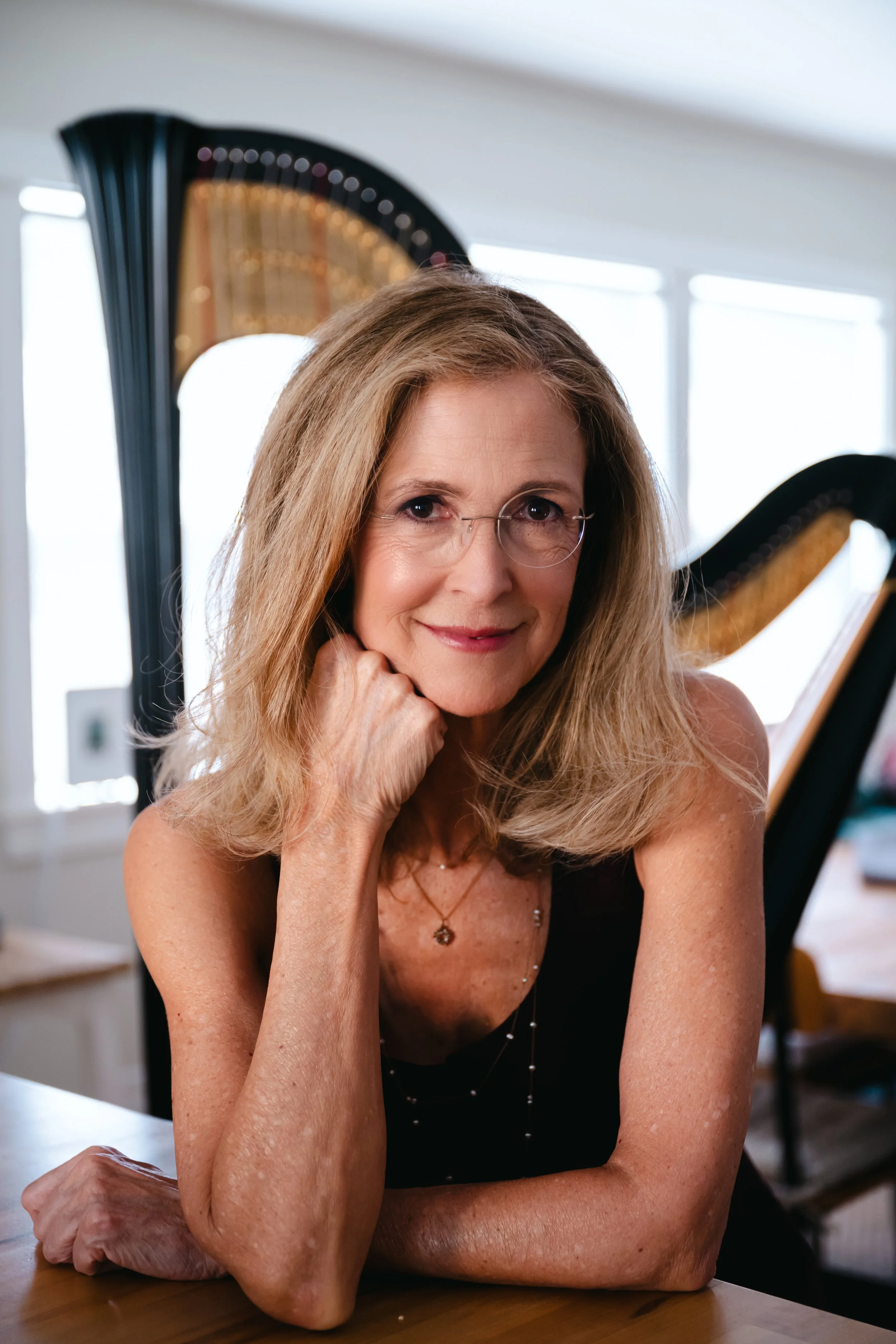A middle-aged woman with blonde hair and glasses, smiling gently, resting her chin on her hand, sitting at a wooden table in a bright room with large windows and a grand piano in the background.