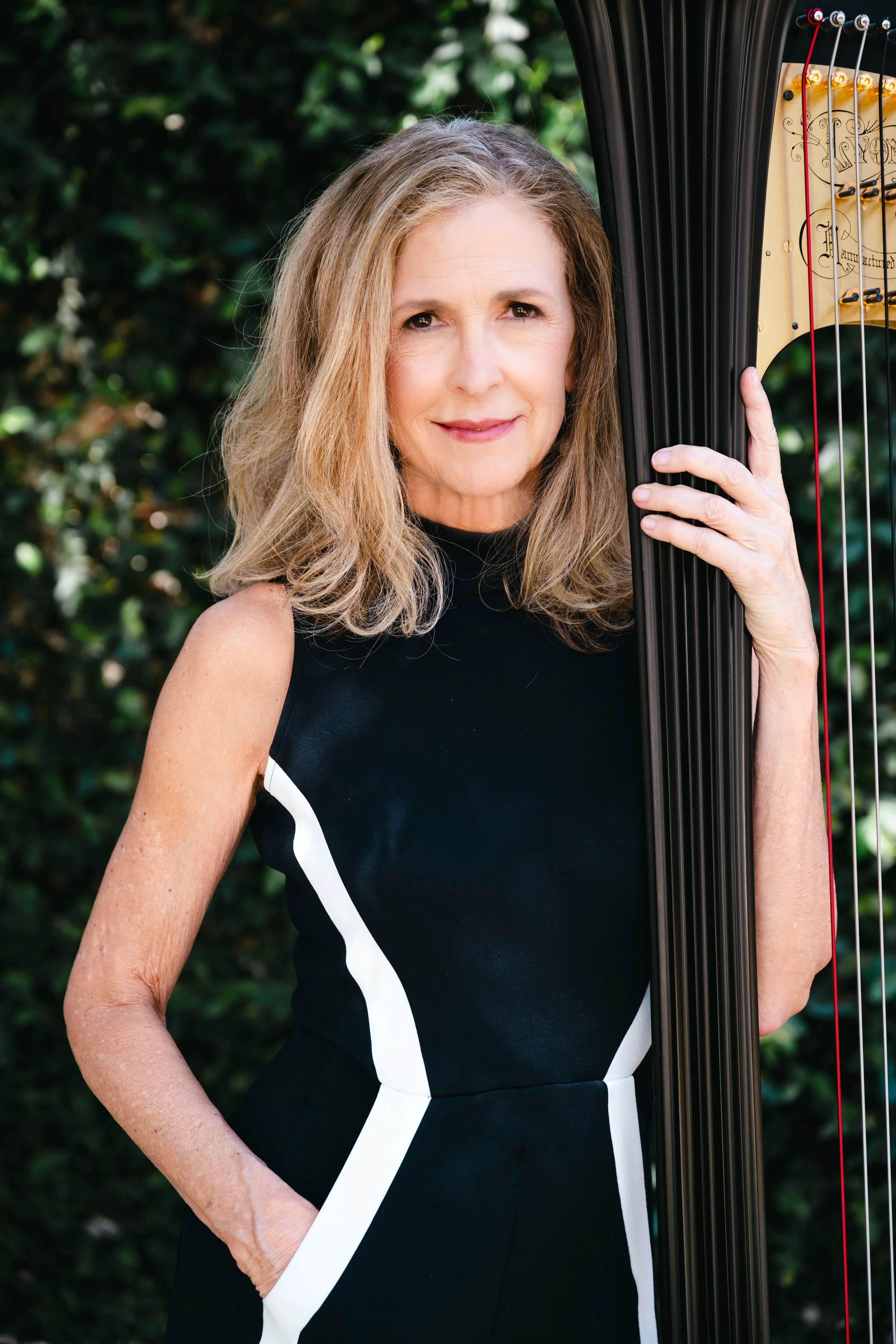 A woman with shoulder-length light brown hair holds a black harp outdoors with a background of green foliage. She is wearing a black sleeveless dress with white accents.