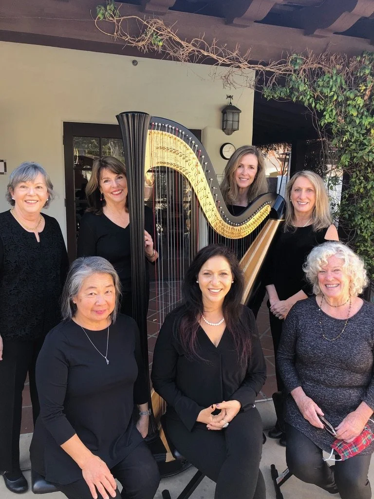 Group of seven women dressed in black, standing and sitting around a harpsichord outside a building, smiling for the camera.