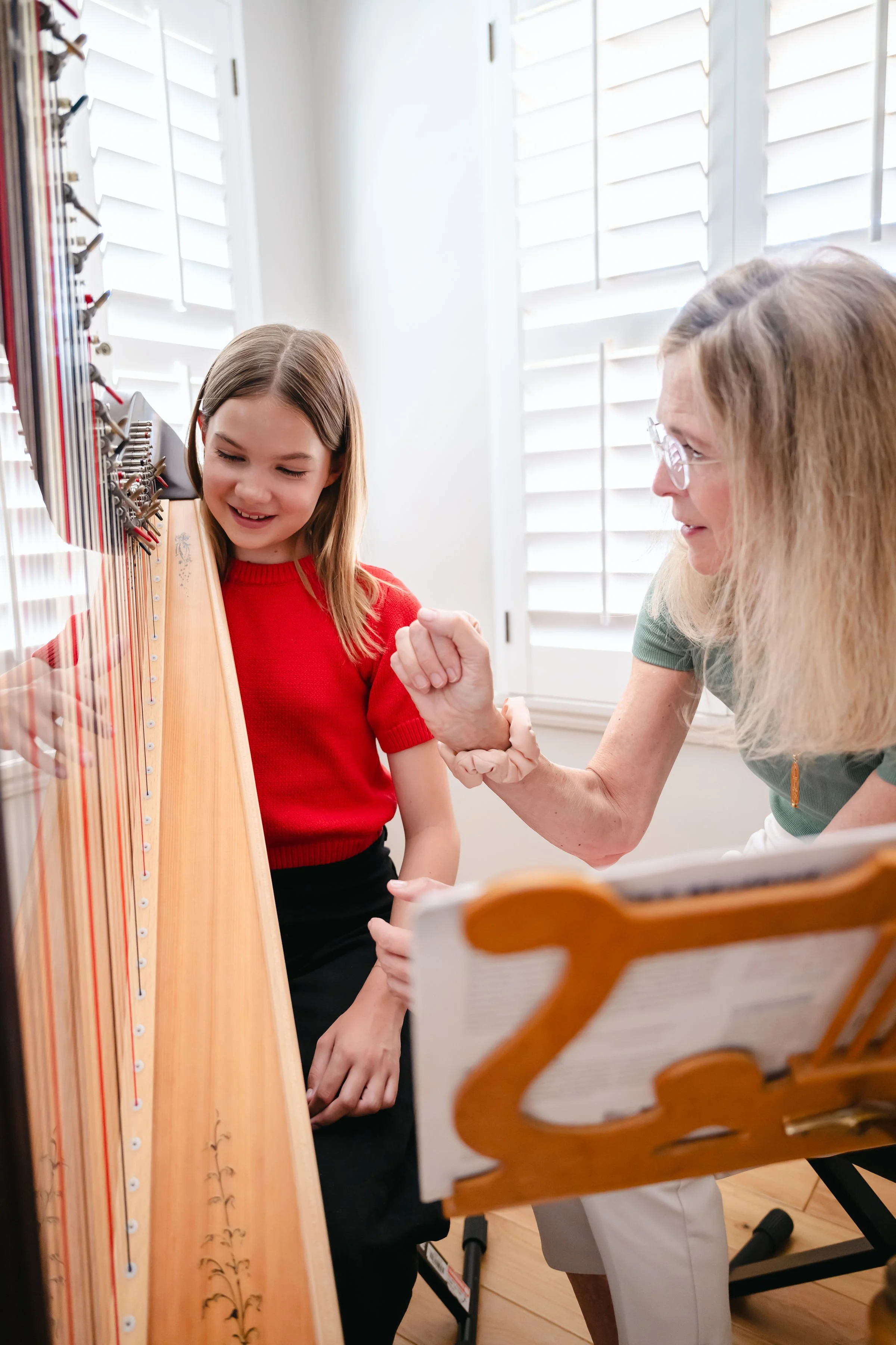 A young girl in a red shirt smiling while playing a hammered dulcimer, with an older woman watching and smiling in a music room with white shutters and wooden floor.