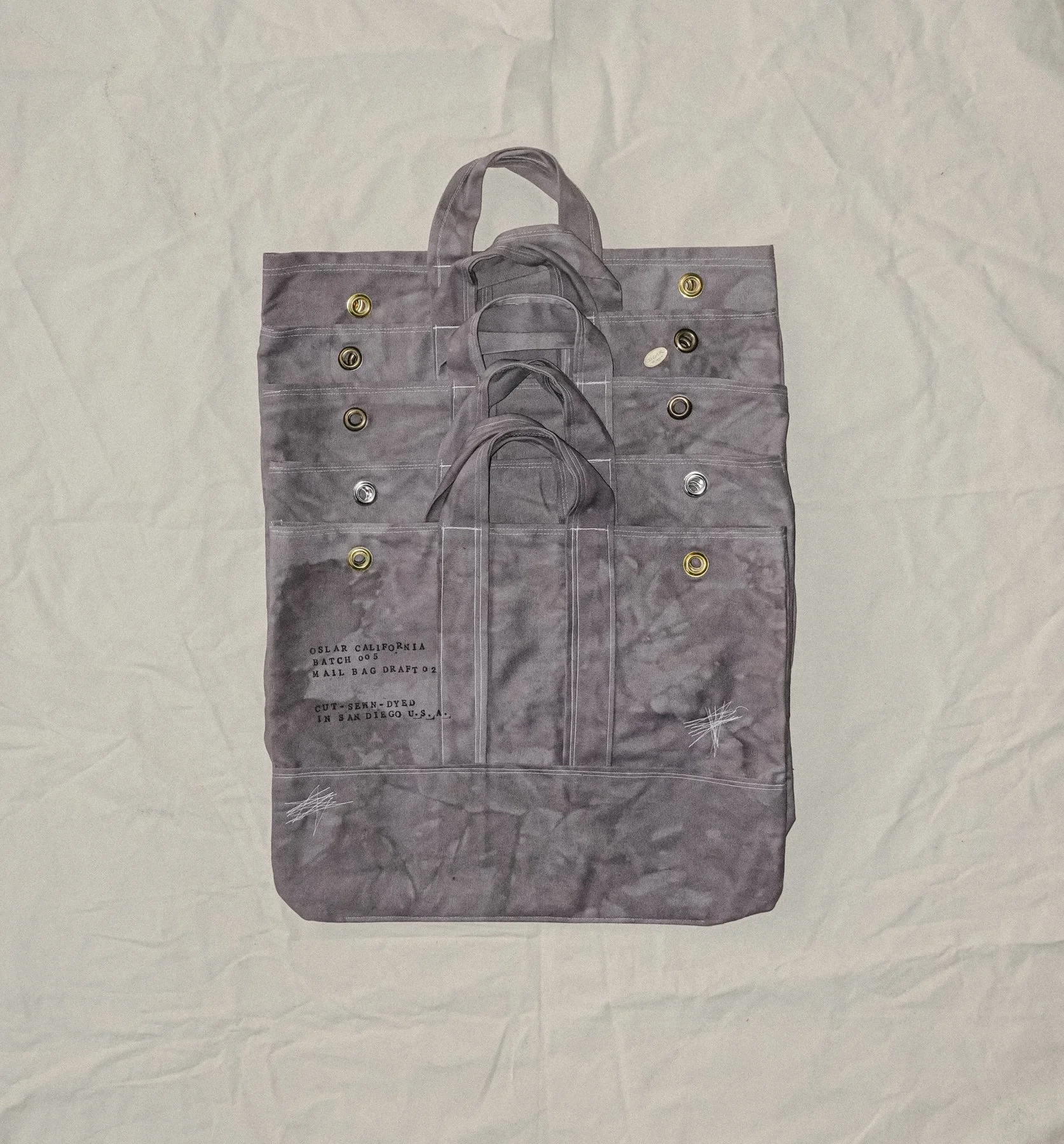 Multiple overlapping gray mail bags with metal eyelets. Text on one bag reads "OSLAR CALIFORNIA BATCH 005 MAIL BAG DRAFT 12 CUT-SEWN- DYED IN SAN DIEGO U.S.A."
