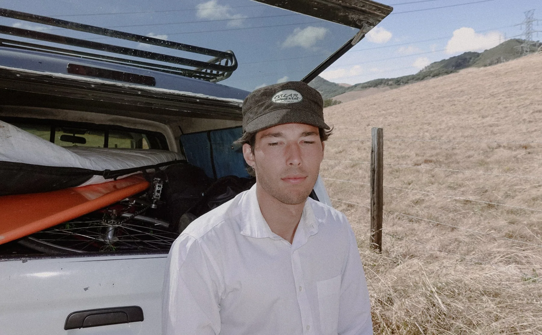 Young man wearing a white shirt and a brown hat standing next to a car with gear and equipment in a rural area on a sunny day.