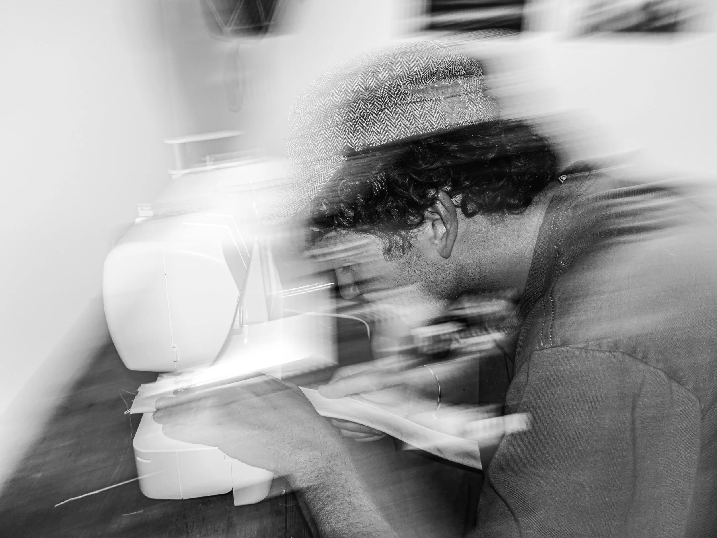 Black and white photo of a person with long curly hair, wearing a cap and board shorts, sitting indoors at a table with a device or paper in front of them.