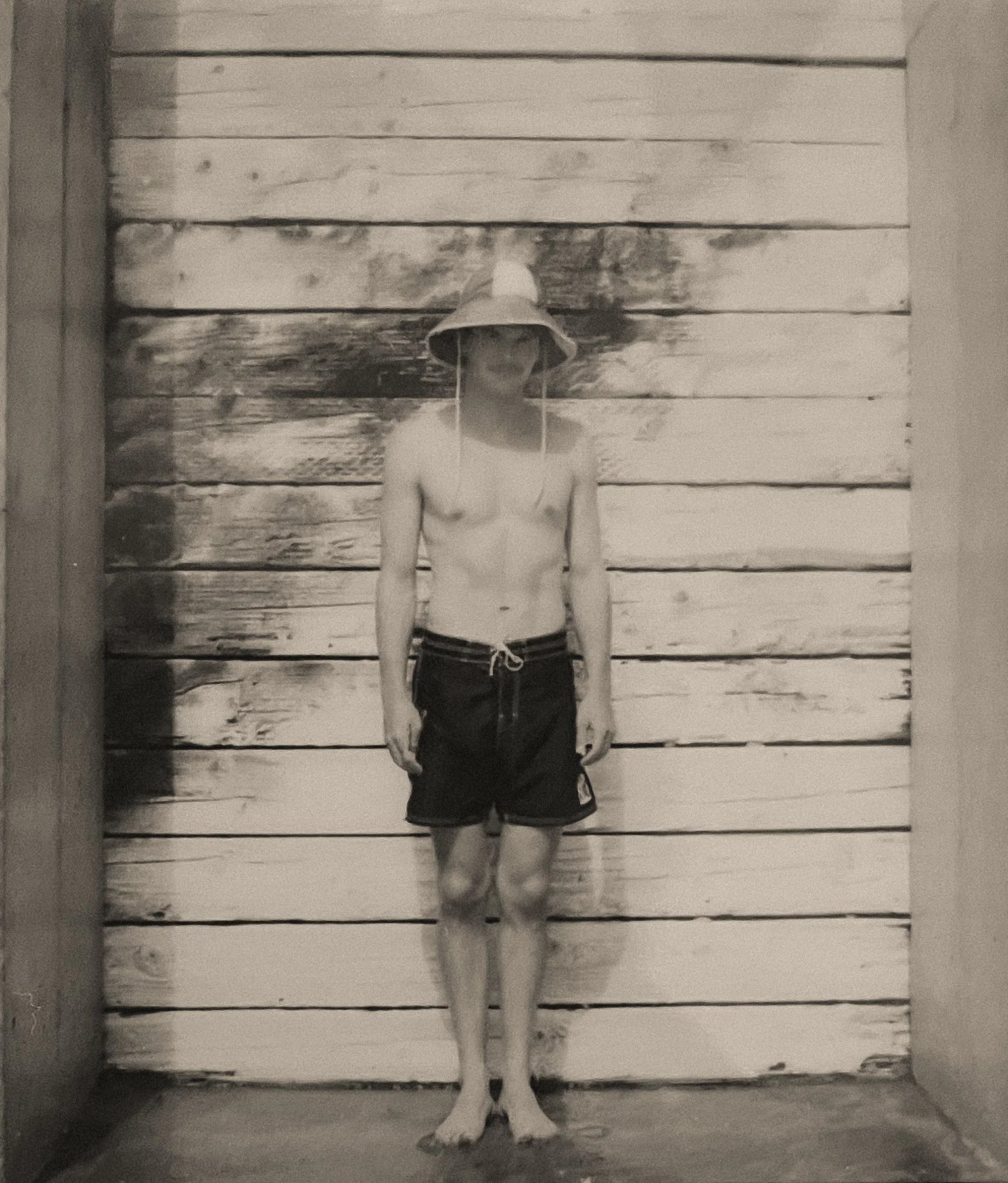 A shirtless man wearing a wide-brimmed hat and shorts is standing in a doorway, with a wooden wall behind him.
