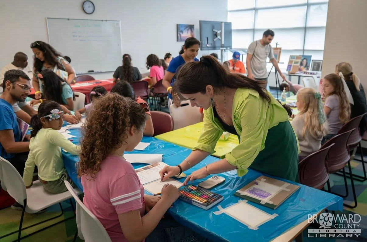 Children and adults participating in arts and crafts activities in a classroom or community room, with supplies on tables and a whiteboard in the background.