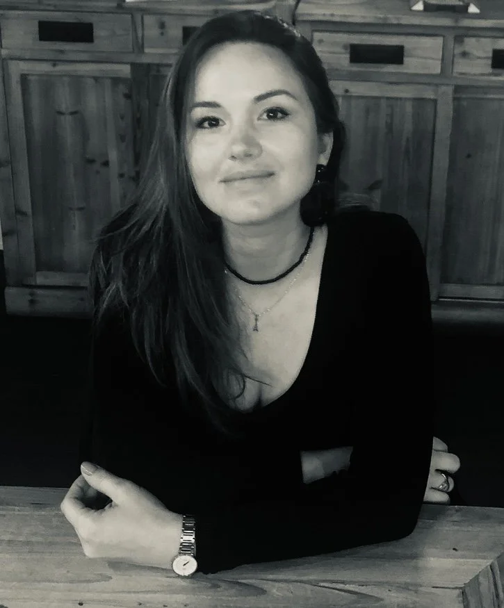 A woman with long hair, wearing a black top, layered necklaces, and a watch, sitting at a wooden table in front of a wooden wall.