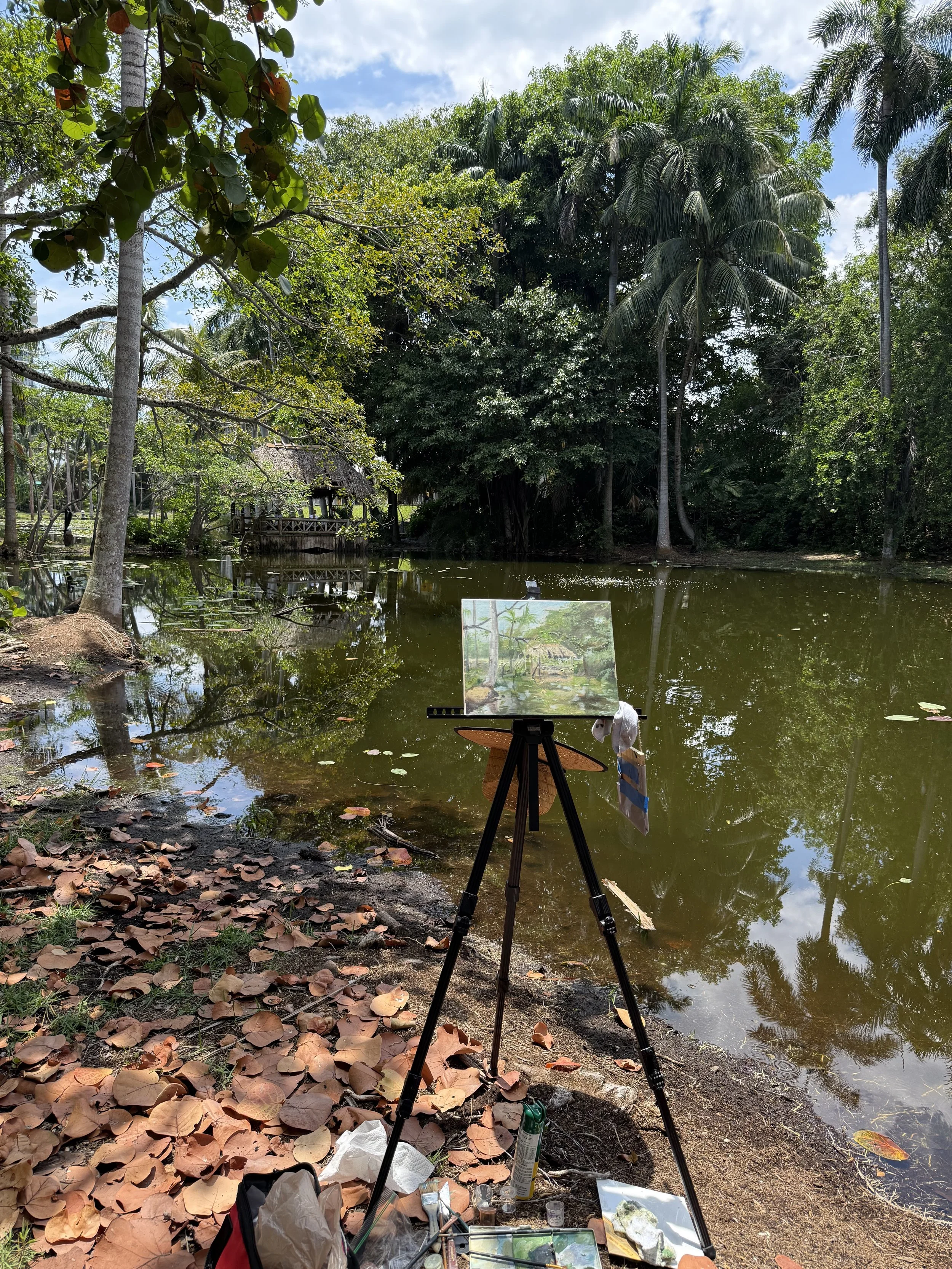 A painting easel holding a canvas depicting a tropical scene with palm trees by a pond, set in a lush forest with many banana trees and a thatched hut in the background. Art supplies and a bag are on the ground, surrounded by fallen leaves.