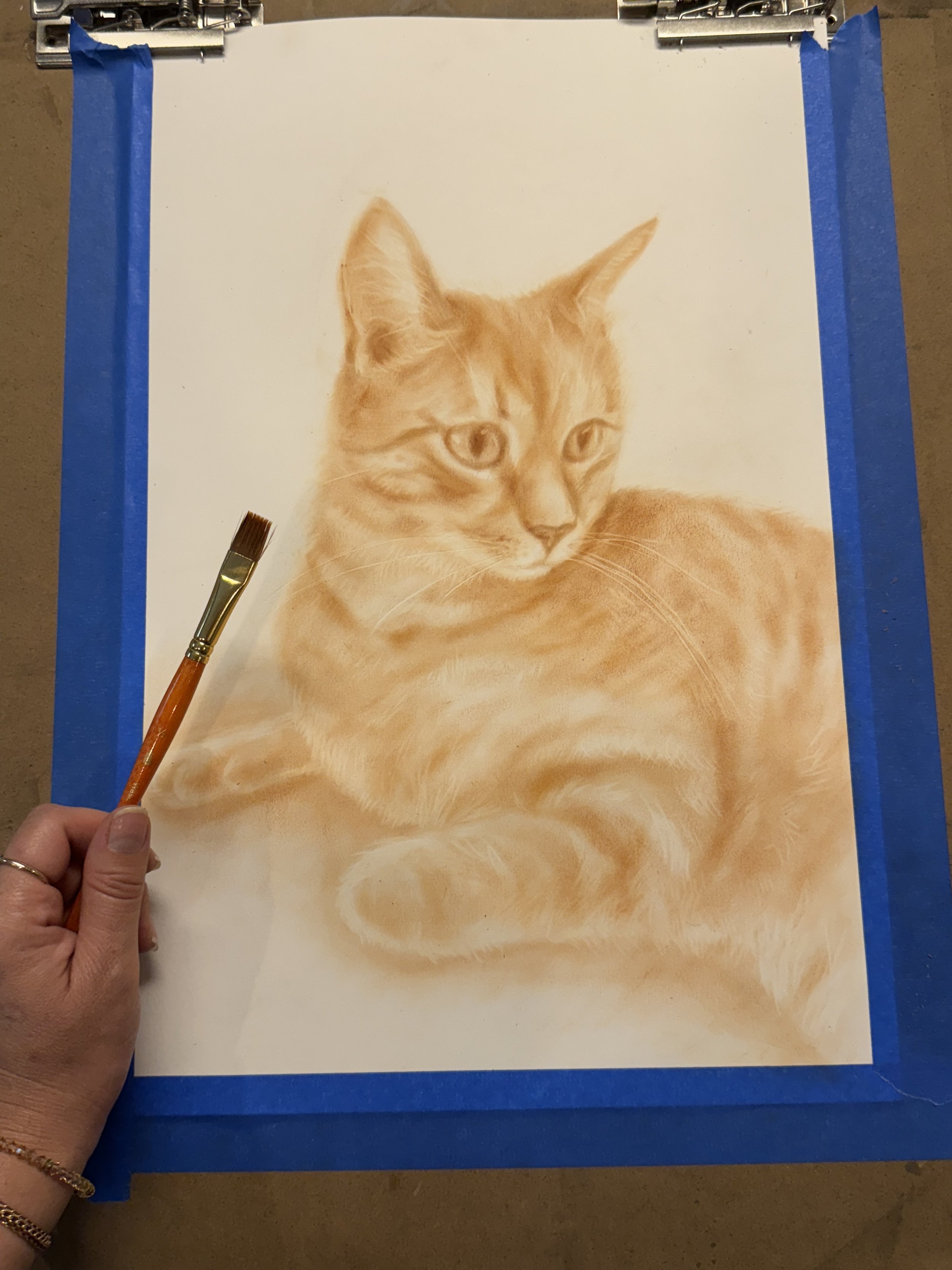 A painting of a cat on a white paper, with a person's hand holding a paintbrush in the lower left corner. Paper is taped to a drawing board