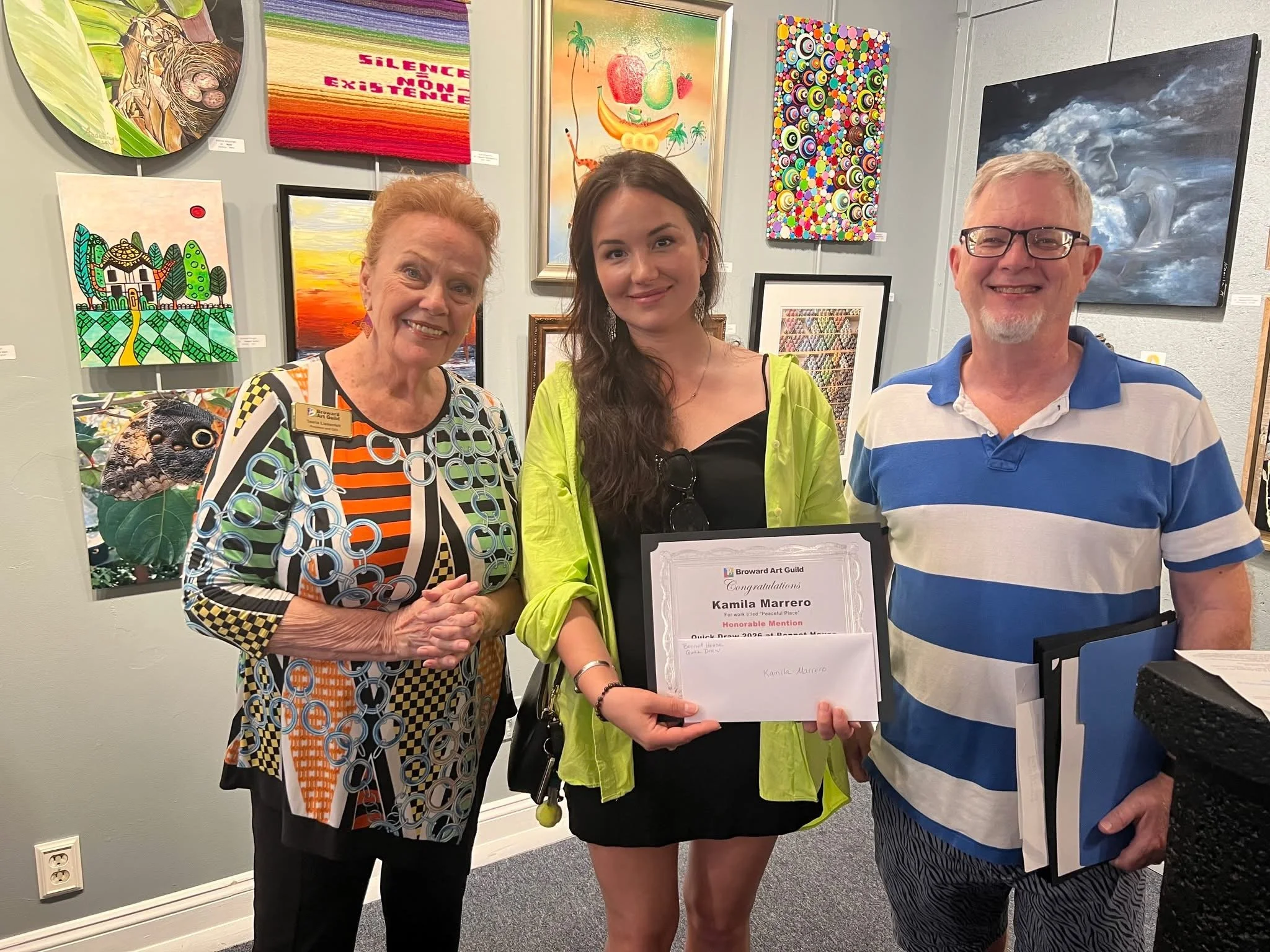 Three people standing in front of colorful artwork at an art gallery. The woman in the middle is holding a congratulatory certificate. The woman on the left has red hair and is wearing a patterned dress, the young woman in the center has long dark hair and is wearing a neon yellow jacket, and the man on the right has short gray hair, glasses, and is wearing a blue and white striped polo shirt.