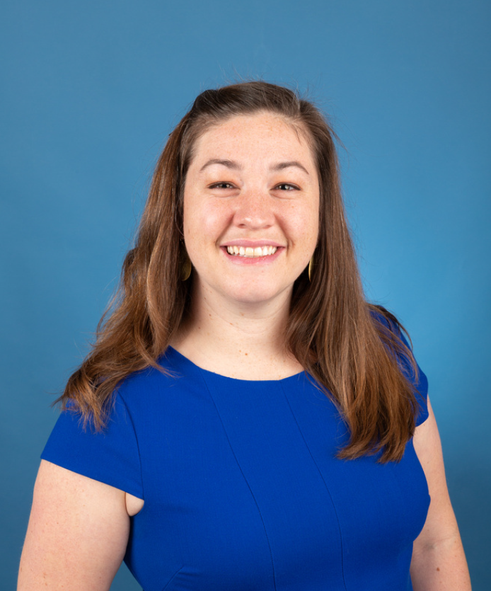 A woman with long brown hair wearing a blue dress, smiling in front of a blue background.