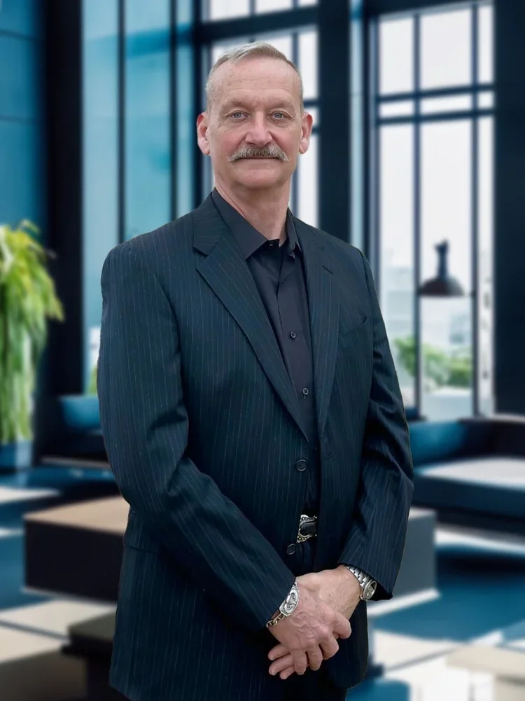 A middle-aged man with a mustache and short hair wearing a dark pinstripe suit standing in a modern office with large windows and a plant in the background.