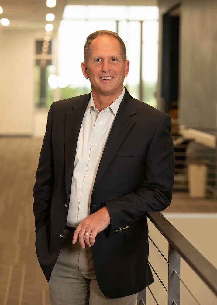 A man in a black suit jacket and white dress shirt standing indoors near a railing, smiling at the camera with a modern office background.