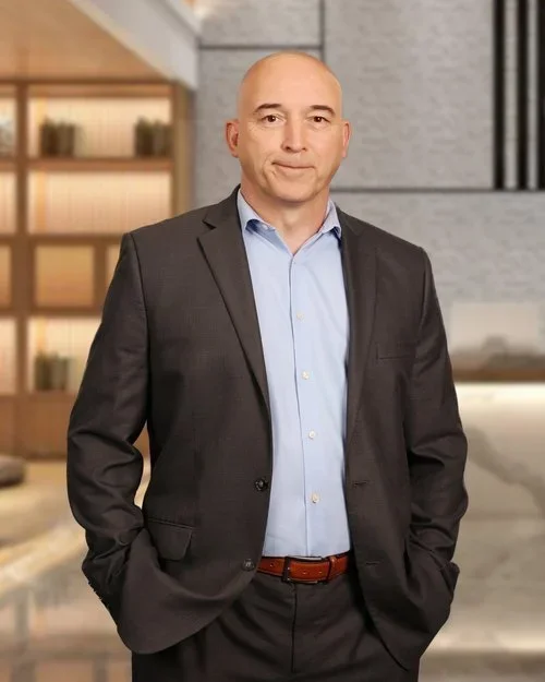 Bald man in a dark suit with a light blue shirt standing in a modern, well-lit interior.