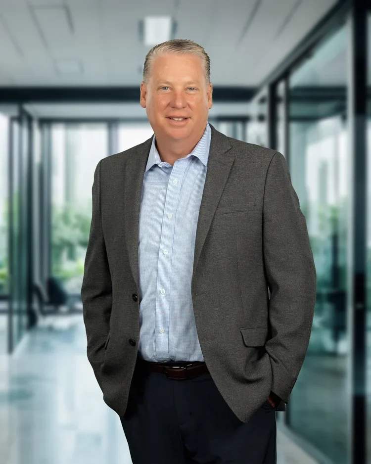 A middle-aged man in a gray suit jacket and light blue dress shirt standing in a modern office building with glass walls.