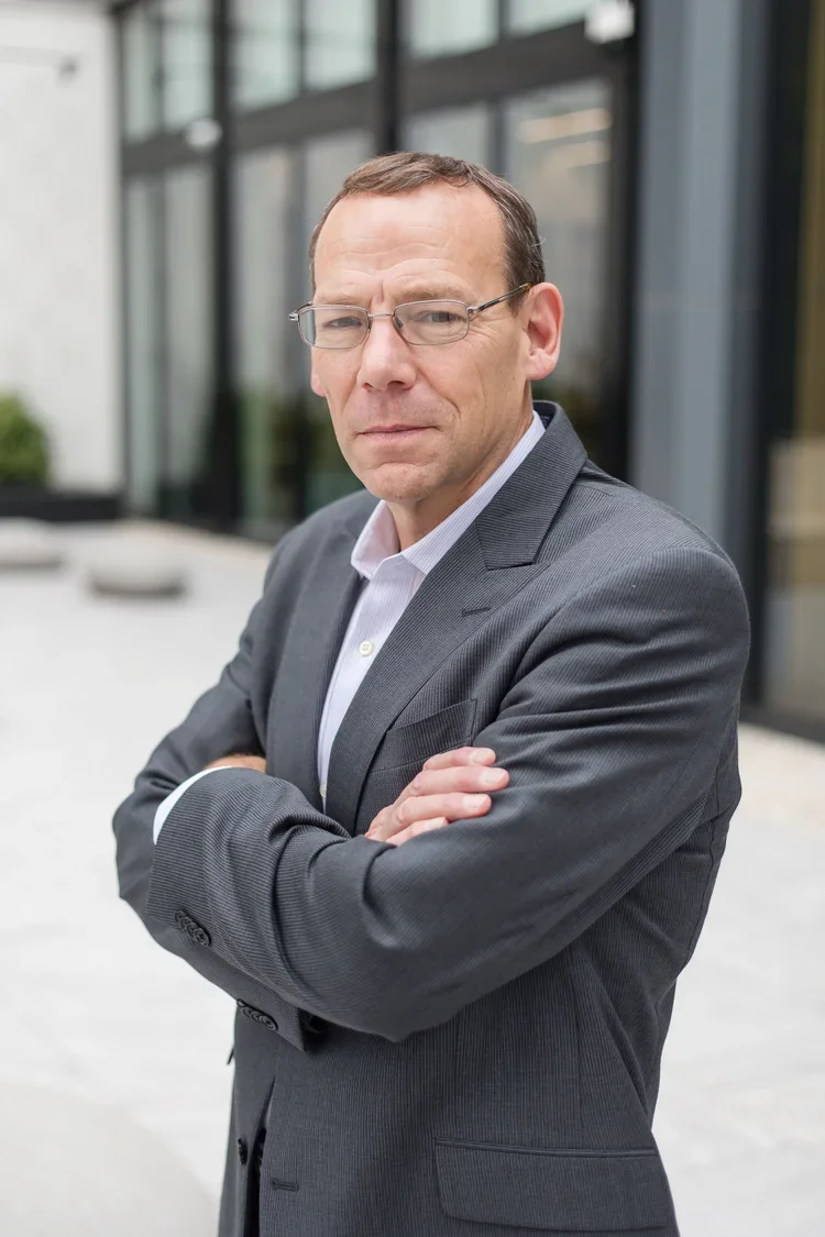 A middle-aged man in a gray suit and glasses standing outside with arms crossed, in front of a modern building with large glass windows.