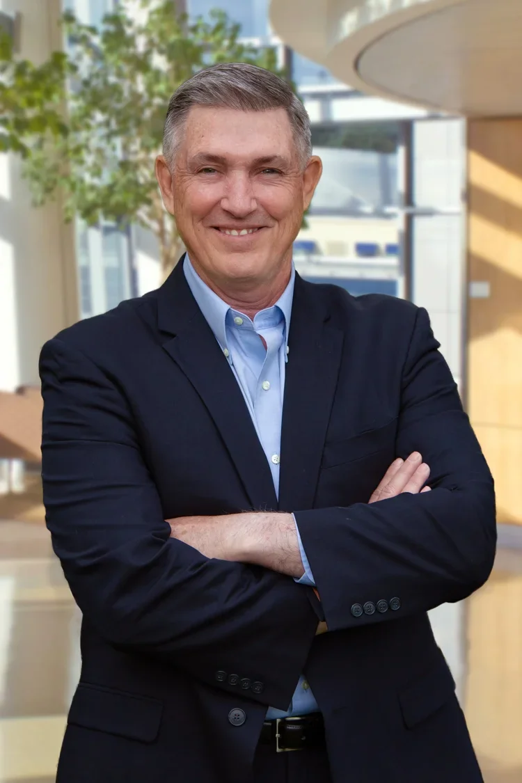 A middle-aged man with short gray hair smiling, standing outdoors with arms crossed, wearing a dark suit jacket and a light blue collared shirt, with a blurred modern office building and greenery in the background.