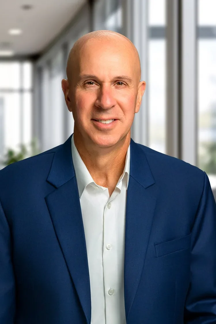A bald man in a blue suit and white shirt smiling in a modern office with large windows.