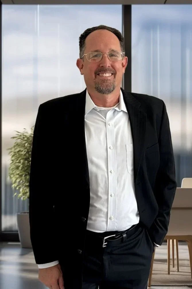 A smiling man in a black suit and white shirt standing in an office with large windows and a plant in the background.