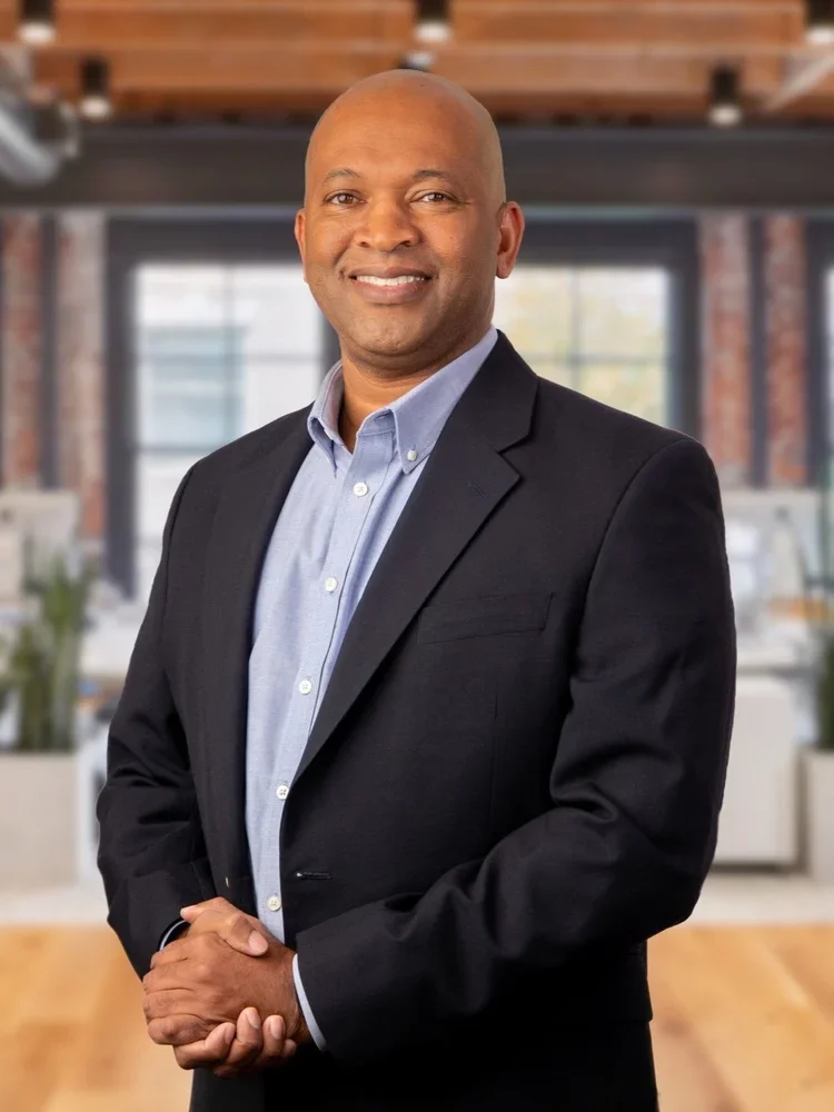 A confident man in a business suit standing in a modern office space with large windows, brick walls, and wooden ceiling beams.