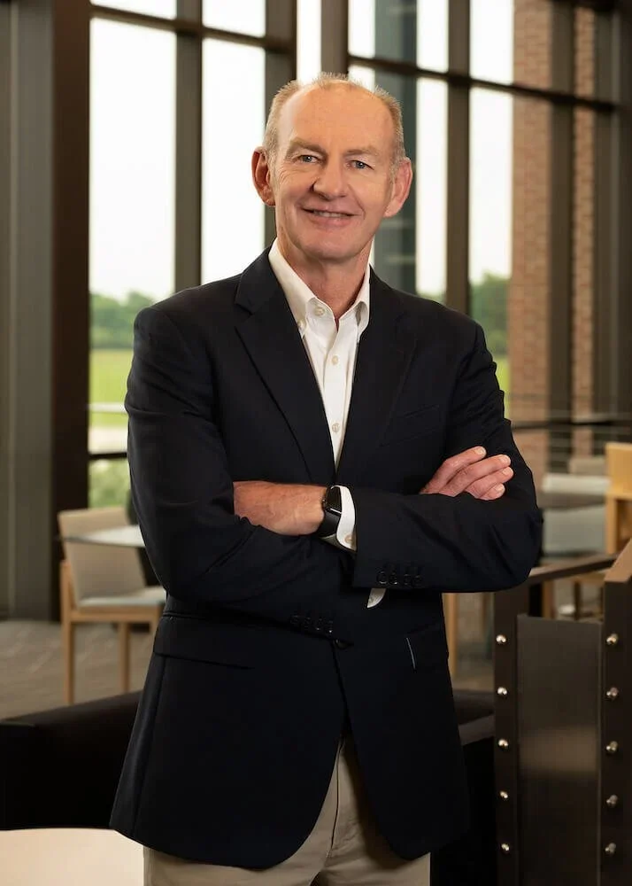 A middle-aged man with light skin, short blonde hair, wearing a black blazer, white shirt, and beige pants, standing with arms crossed inside a modern office or conference room with large windows and a green outdoor view.