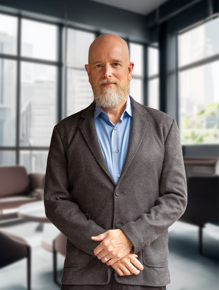 A mature man with a bald head and white beard standing in a modern office with large windows, wearing a gray blazer over a blue shirt.