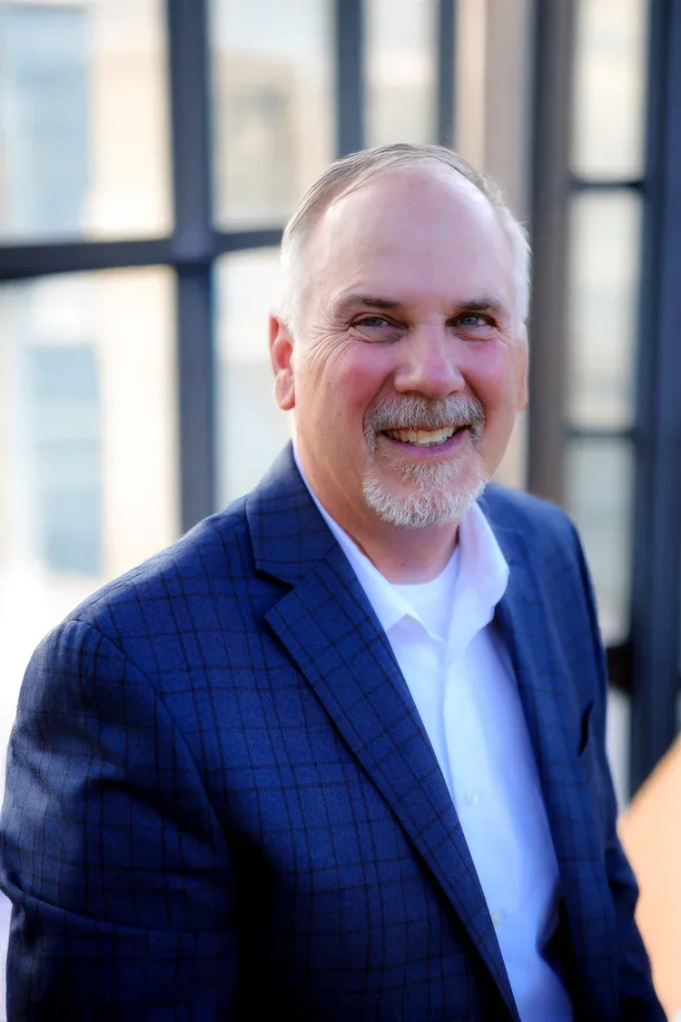 Professional man smiling, wearing a blue checked blazer and white shirt, standing indoors near large windows.