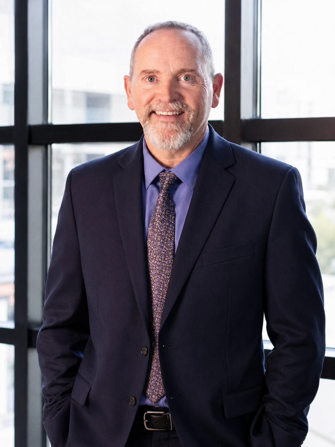 A smiling man with gray hair and beard, wearing a dark suit, blue shirt, and patterned tie, standing in front of a window with a cityscape background.