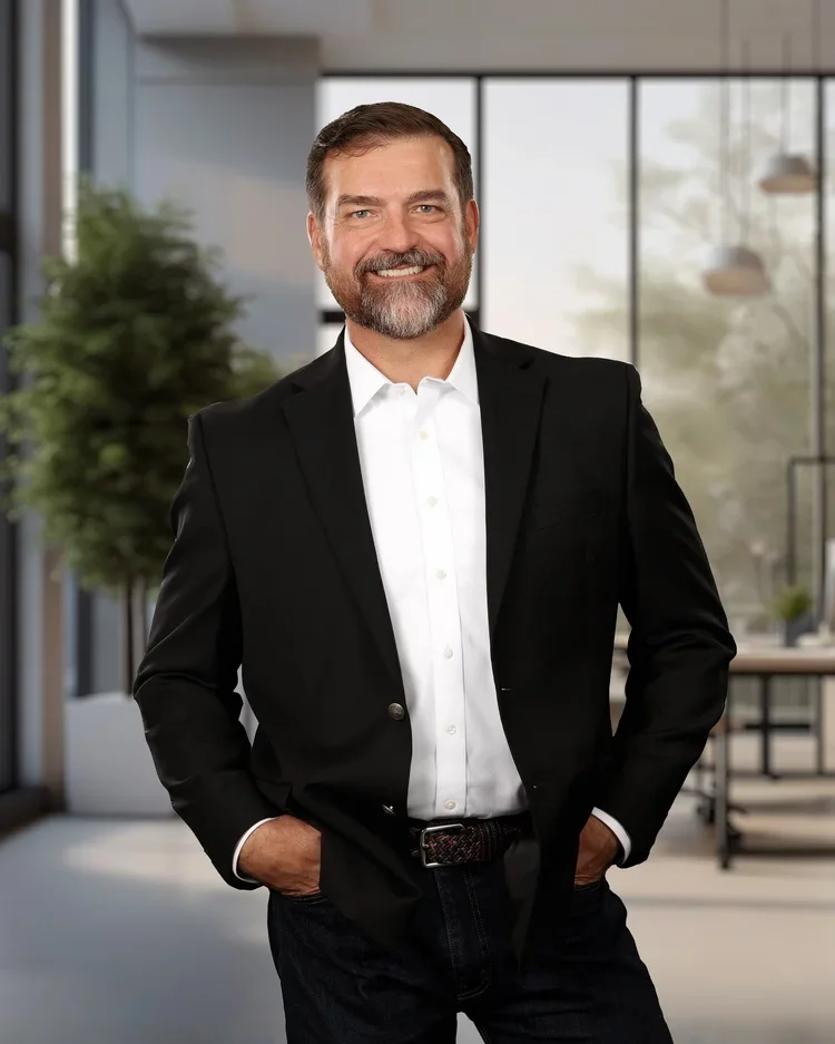 A man with a beard and short hair, wearing a black suit jacket, white dress shirt, and jeans, standing in a modern office with large windows and plants.
