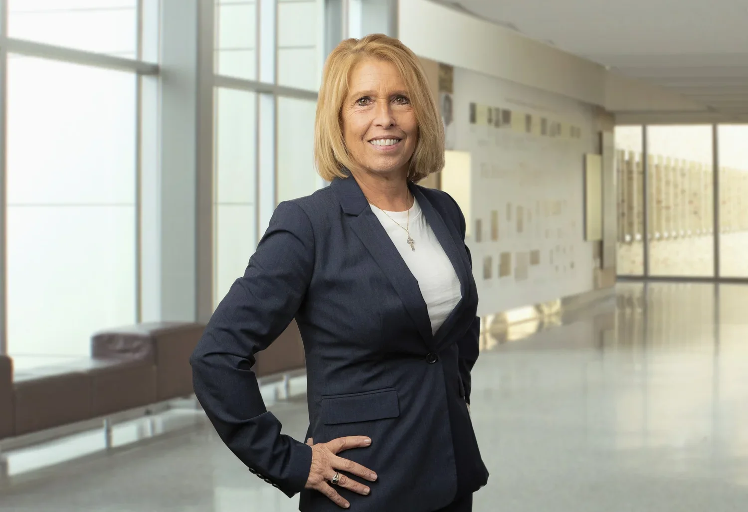 A confident woman with shoulder-length blonde hair, wearing a navy blue blazer over a white shirt, standing with one hand on her hip in a modern, well-lit corridor with large windows.