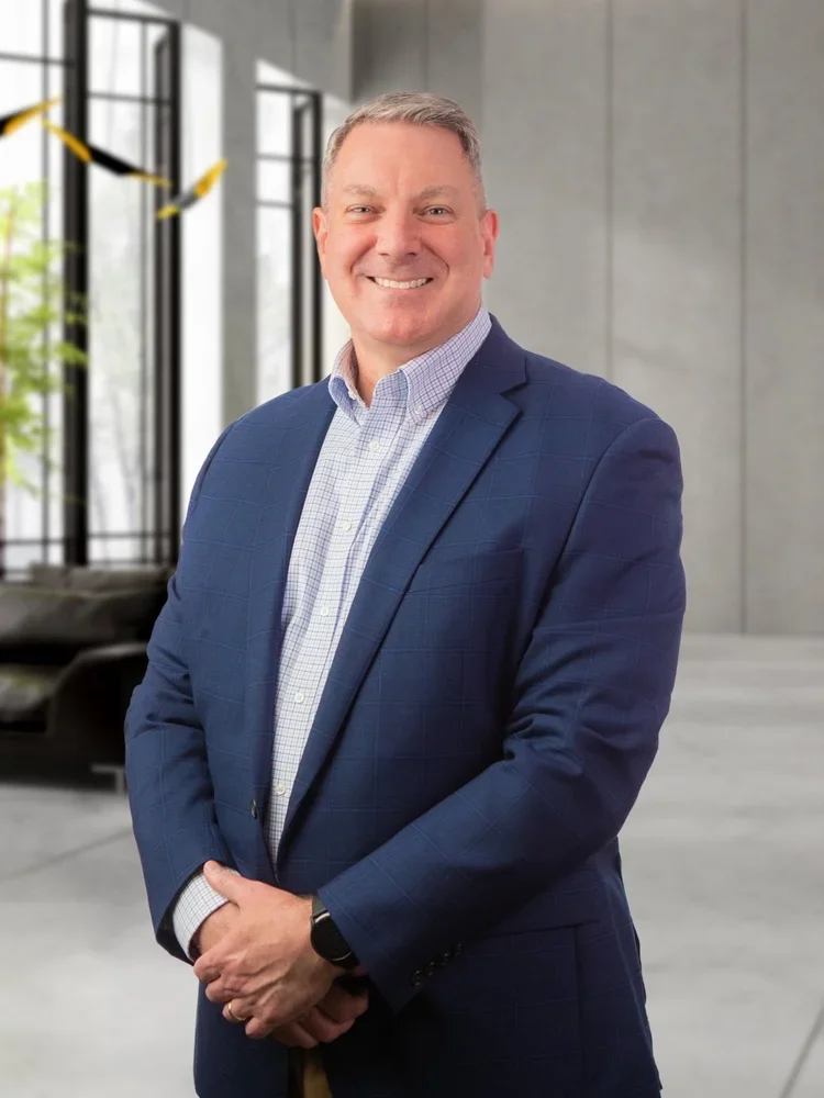 A professional man in a blue suit and checkered shirt standing in a modern office space with large windows and minimal decor, smiling at the camera.